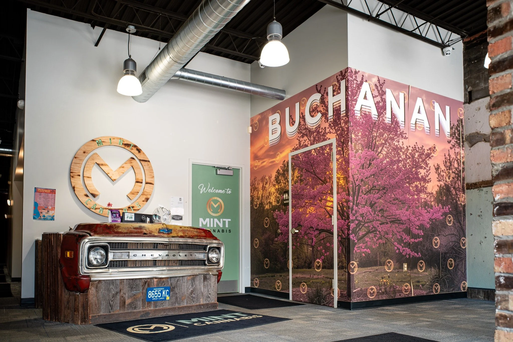 Indoor space with industrial ceiling, ductwork, and lighting. Decorative wall panel features a pink flowering tree scene with large 3D letters spelling 'BUCHANAN'. A vintage Chevrolet truck front is displayed on a wooden platform with a Michigan lice
