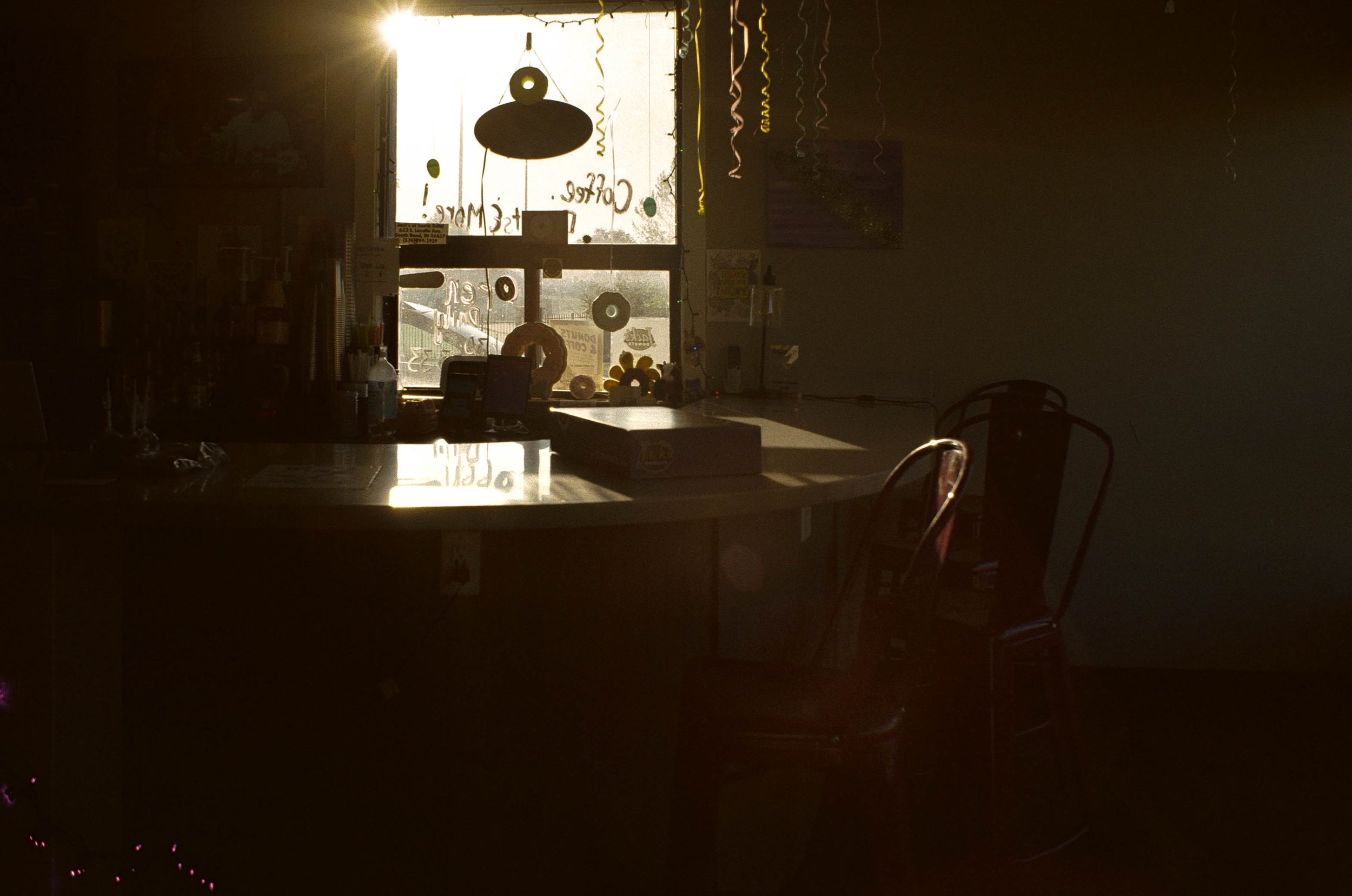 A dimly lit indoor space with a bright window in the background. The window has holiday decorations and writing on it, with some objects and furniture partially visible in the low light.