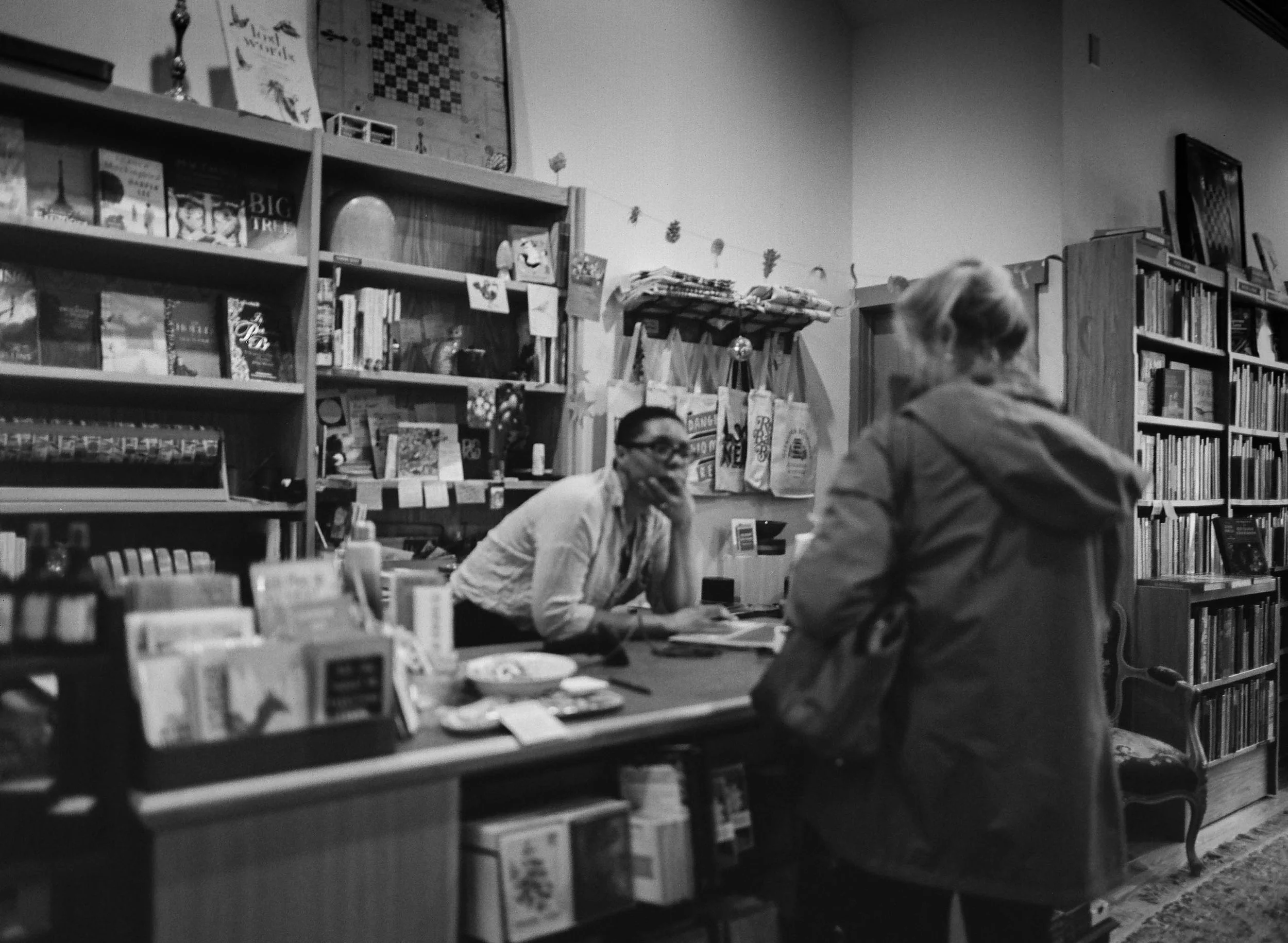 Black and white photo of a bookstore or library with shelves filled with books. Two people are engaged in conversation, one facing the camera and the other with their back to it, seated at a table.