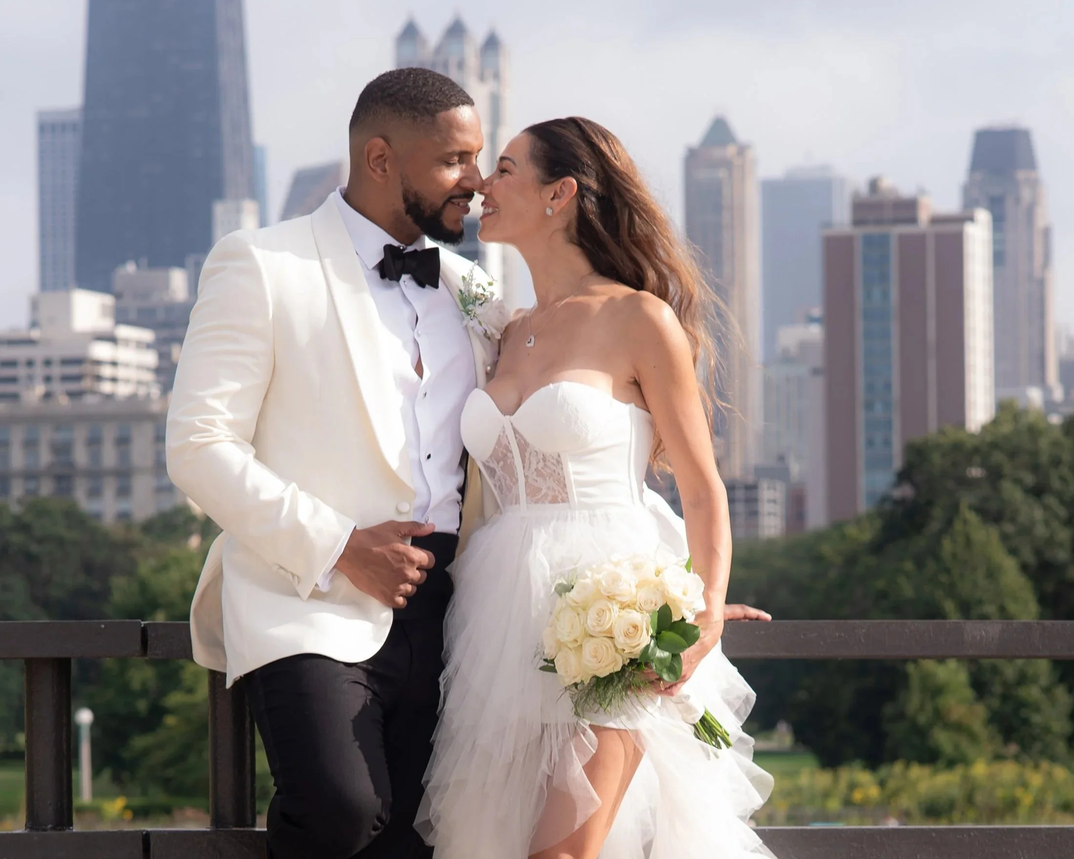 A newlywed couple standing close together on a bridge with a city skyline in the background. The bride is in a strapless white wedding dress with lace details, holding a bouquet of white roses, and wearing white high-heeled sandals. The groom is in a