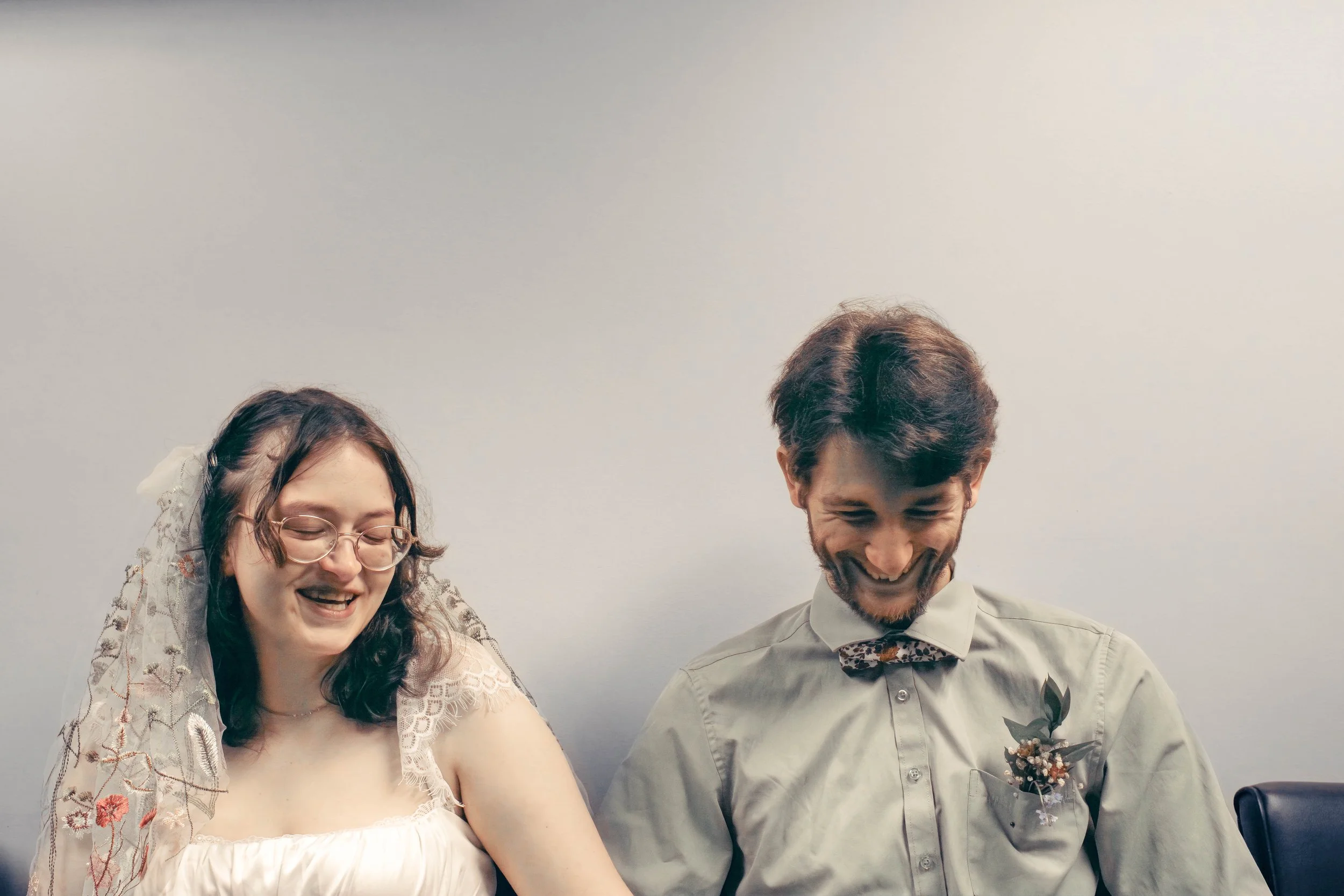 A woman and a man are smiling and looking down, possibly at something they are doing together. The woman is wearing glasses and a lace dress, and the man is dressed in a light-colored shirt with a bow tie. They appear to be happy and enjoying the mom