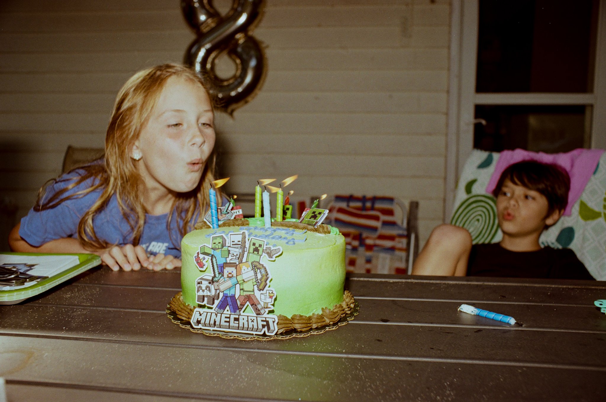 A young girl blowing out candles on a Minecraft-themed birthday cake with another girl sitting nearby, in a room with wooden walls and a window.