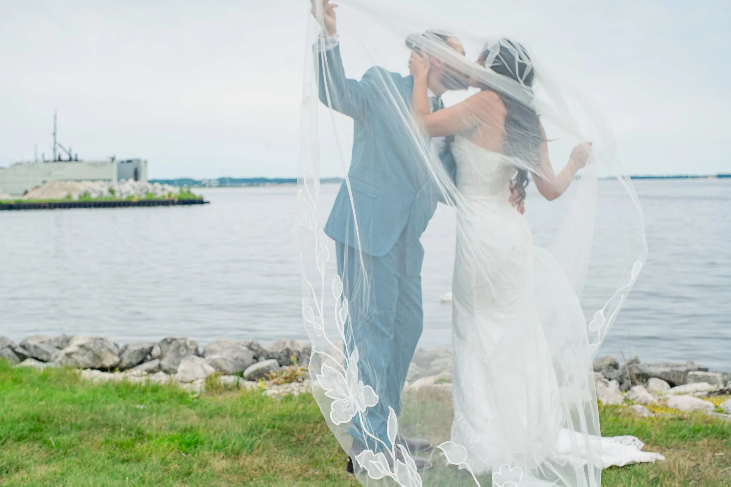 A couple dressed in wedding attire, the groom in a blue suit and the bride in a white dress, embrace under a transparent umbrella by the water with a ship in the background.