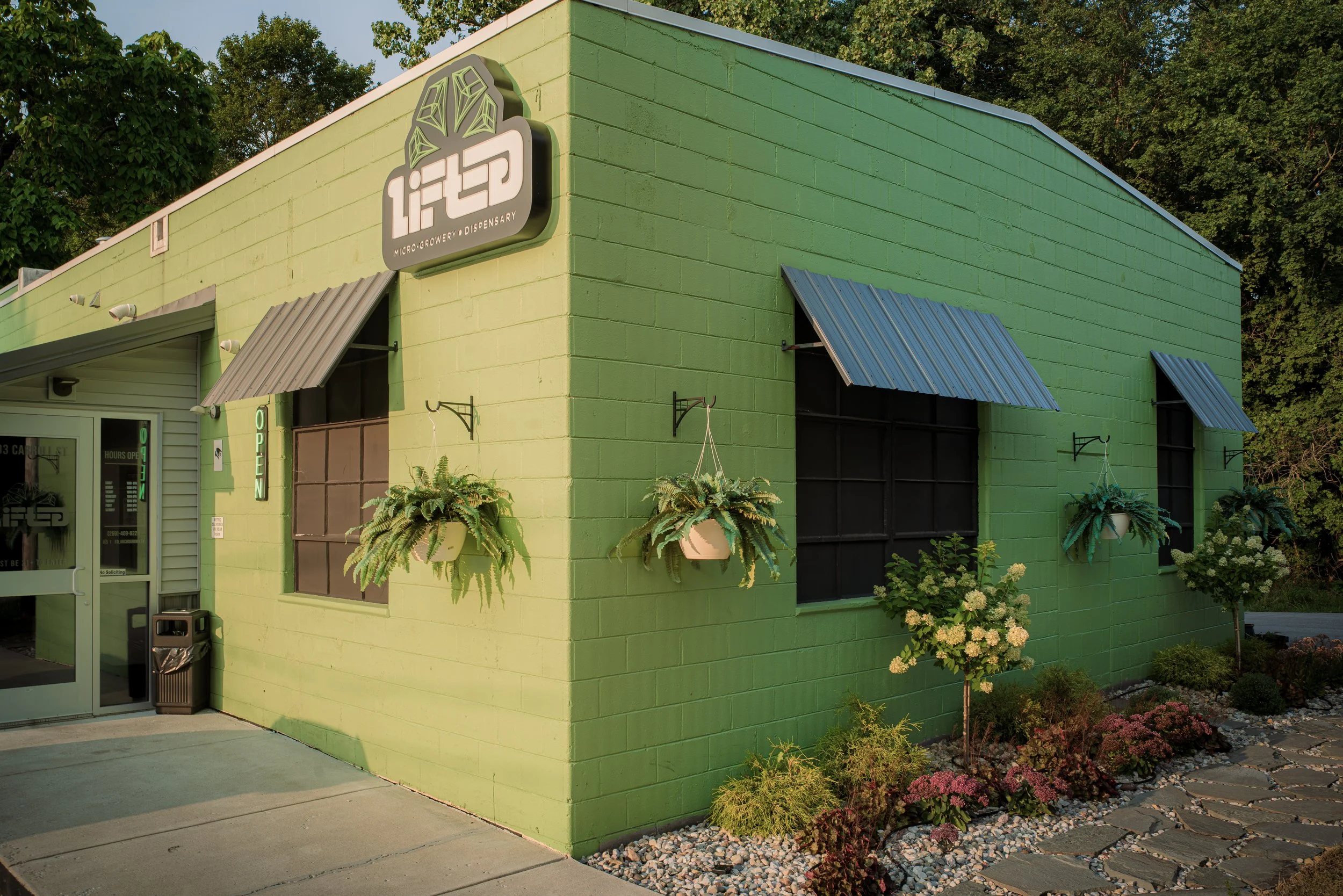 A bright green building with the sign 'LITD Micro-Growery & Dispensary' on the side, featuring plants hanging outside and purple awnings over the windows. The building is surrounded by greenery and small landscaped garden with flowers.