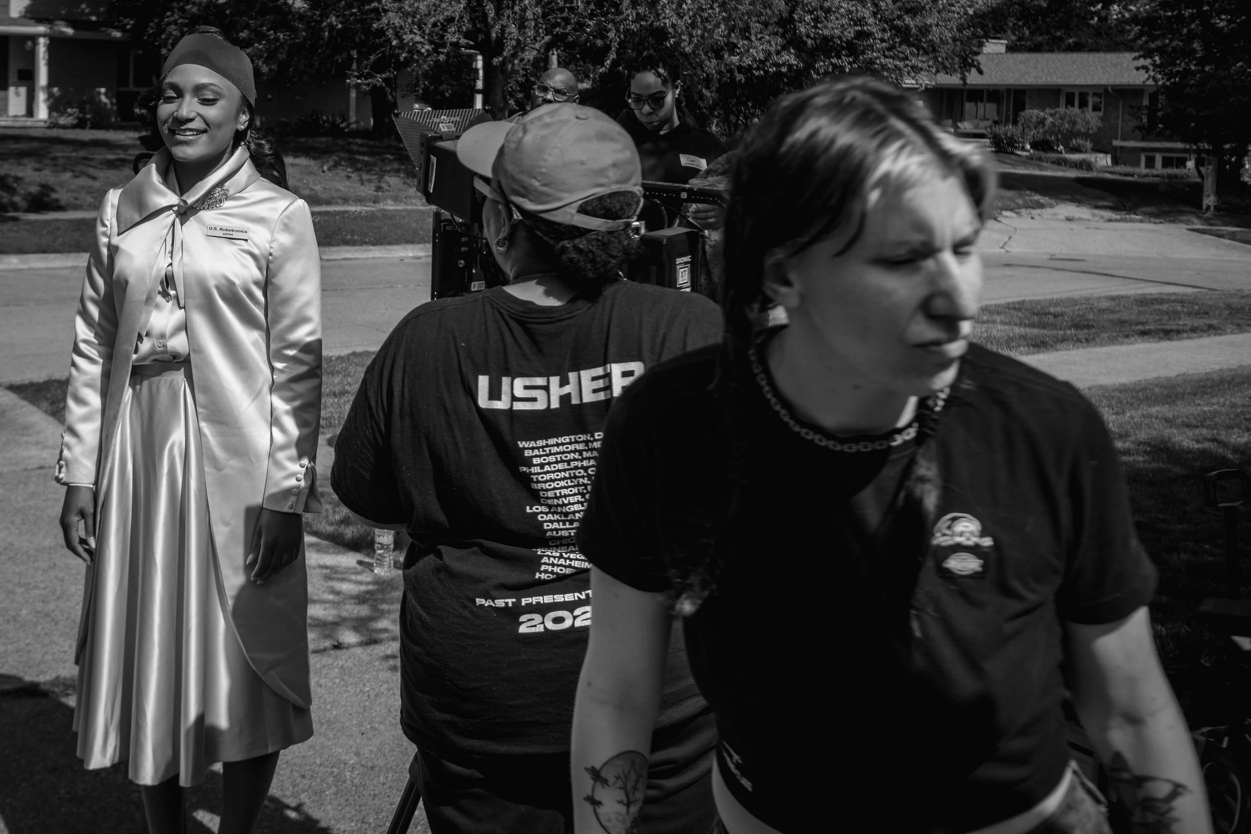 A group of people gathered outdoors, with one woman dressed as Rosie the Riveter, smiling with eyes closed, wearing a satin-like jacket and headscarf. Several people are seen in the background, some facing away from the camera, with trees and residen