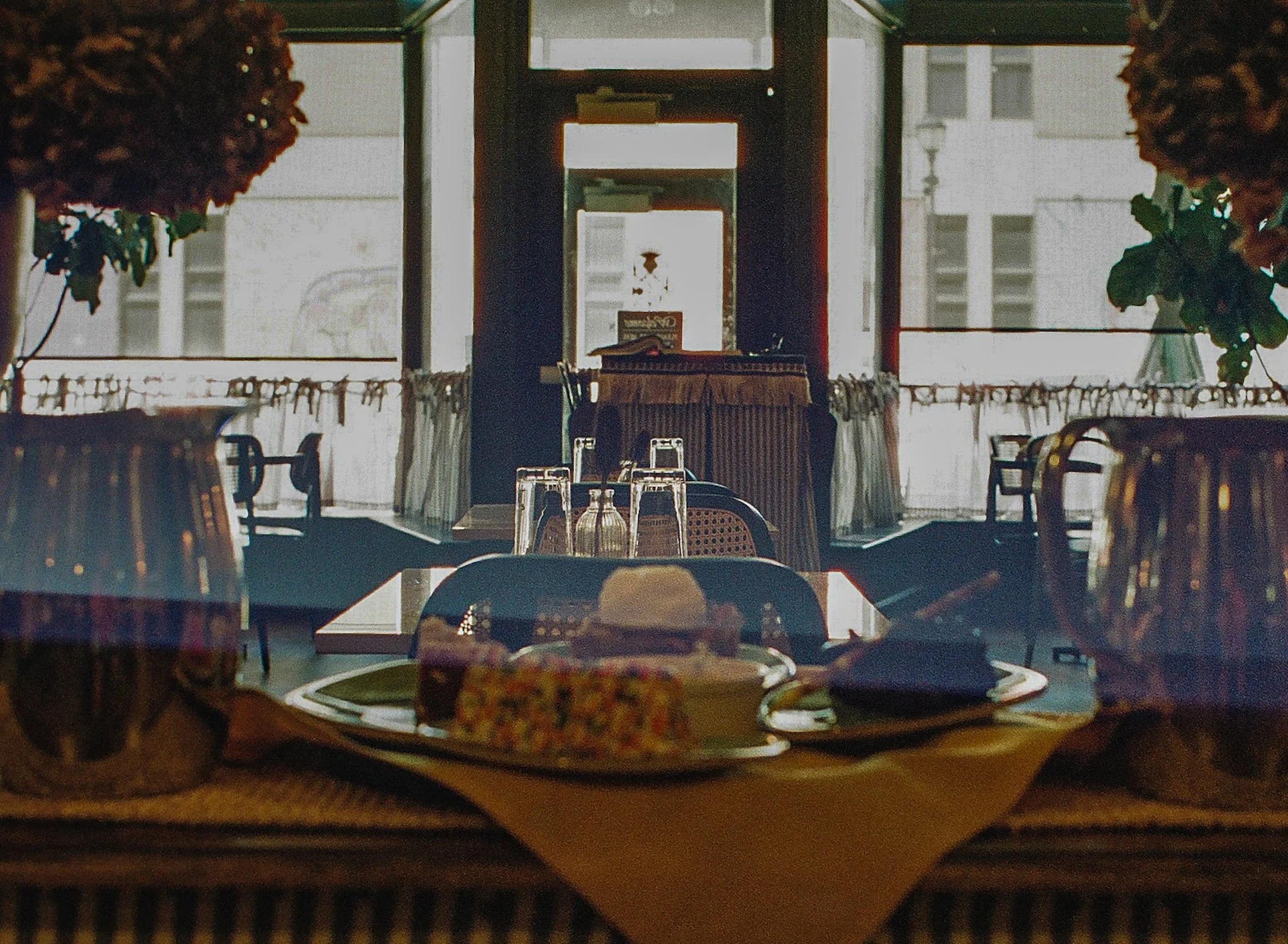 The image reveals the interior of a restaurant viewed through a glass window with reflections. Inside, there are tables and chairs arranged in a dining area. The back of the restaurant features large floor-to-ceiling windows, allowing natural light t