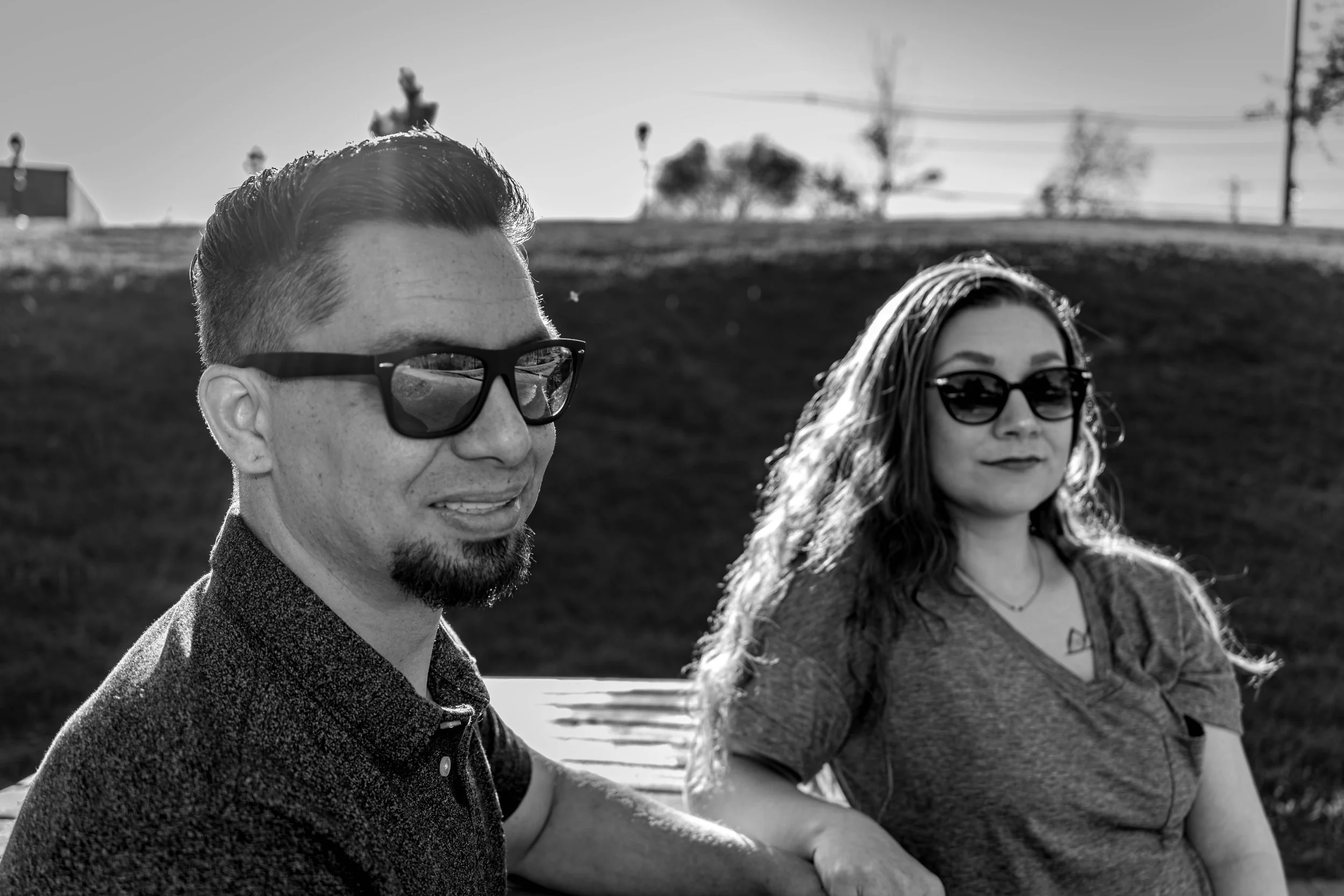 Black and white photo of a man with short hair, beard, and sunglasses, and a woman with long hair and sunglasses, sitting outdoors by the water with trees and power lines in the background.
