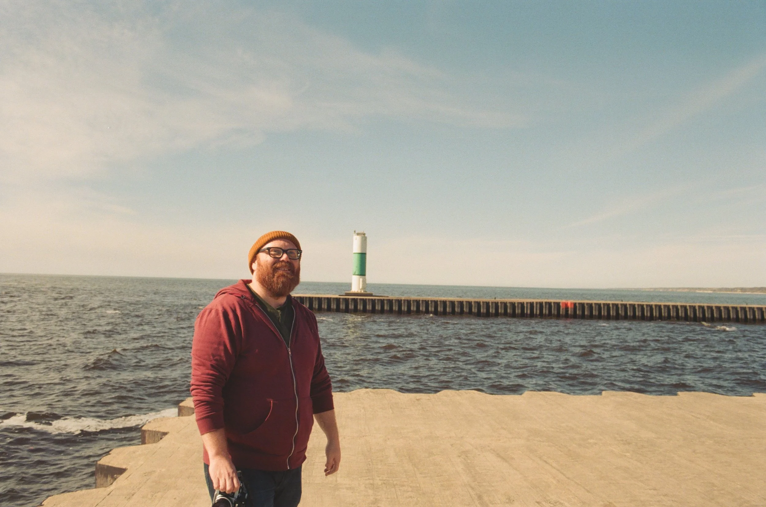 A man with red hair, beard, and glasses wearing a brown knit cap and a maroon hoodie stands on a seaside dock, with a lighthouse and ocean in the background.