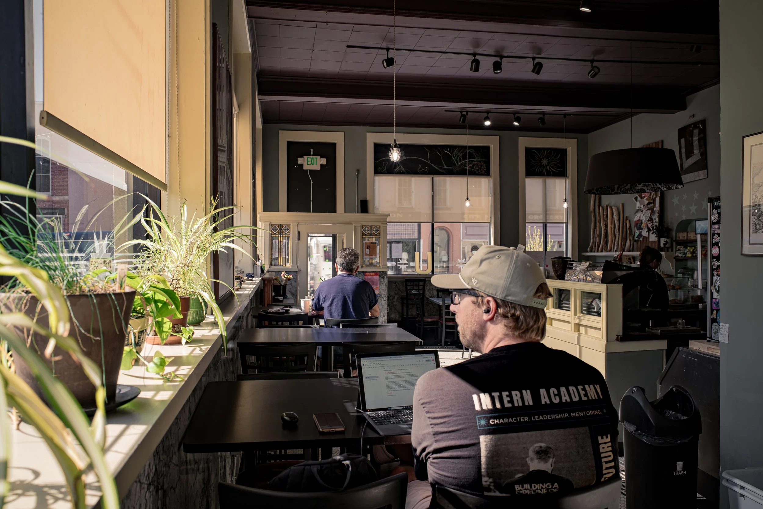 Interior of a cafe with large windows, green plants on a windowsill, and multiple pendant lights hanging from a dark wooden ceiling. Customers are seated at tables, including a man wearing a hat and a t-shirt with “Future” printed on the back. The at