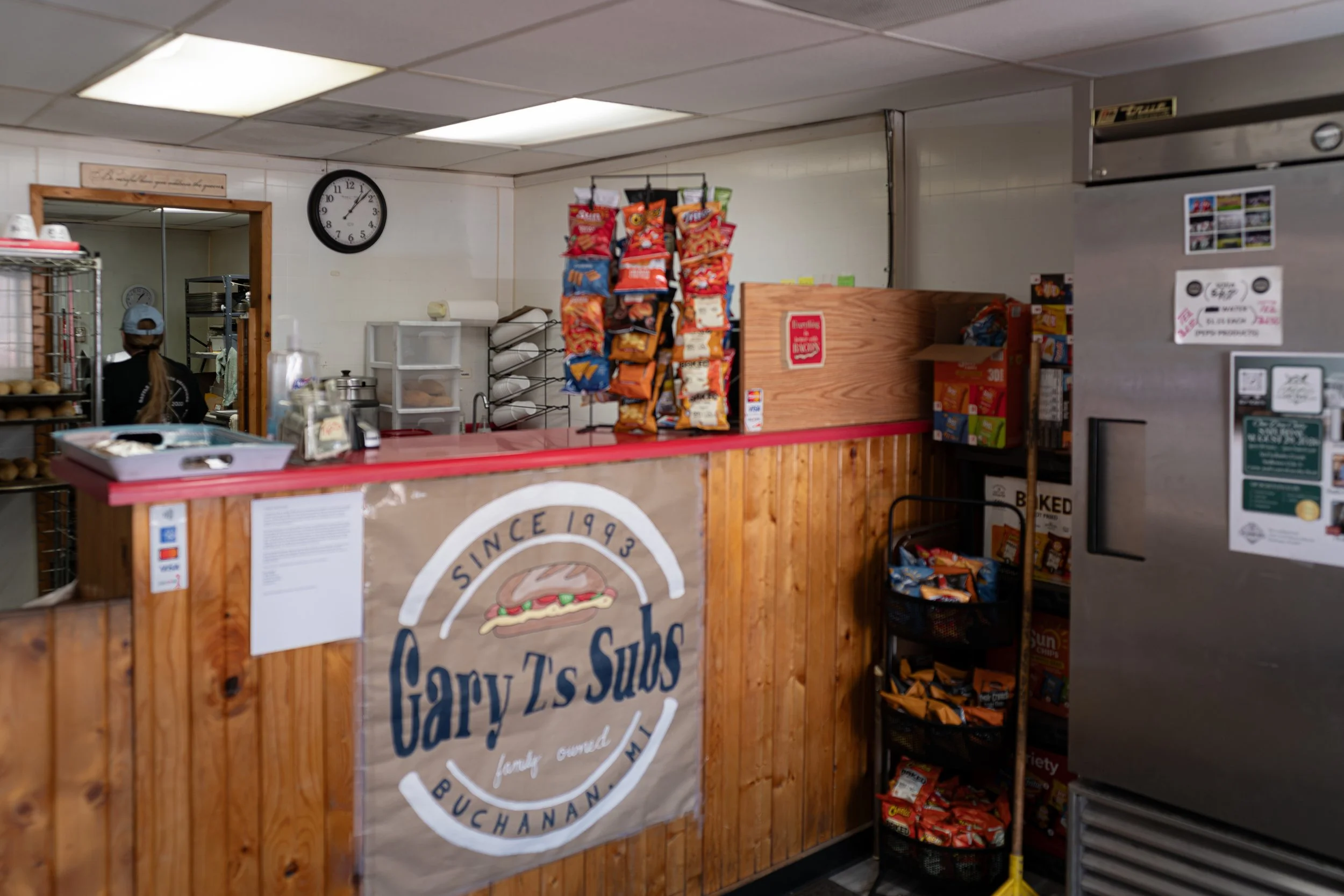 A small convenience store counter with a pizza sign, snack racks with chips hanging above, a clock on the wall, and shelves with snacks and other items.