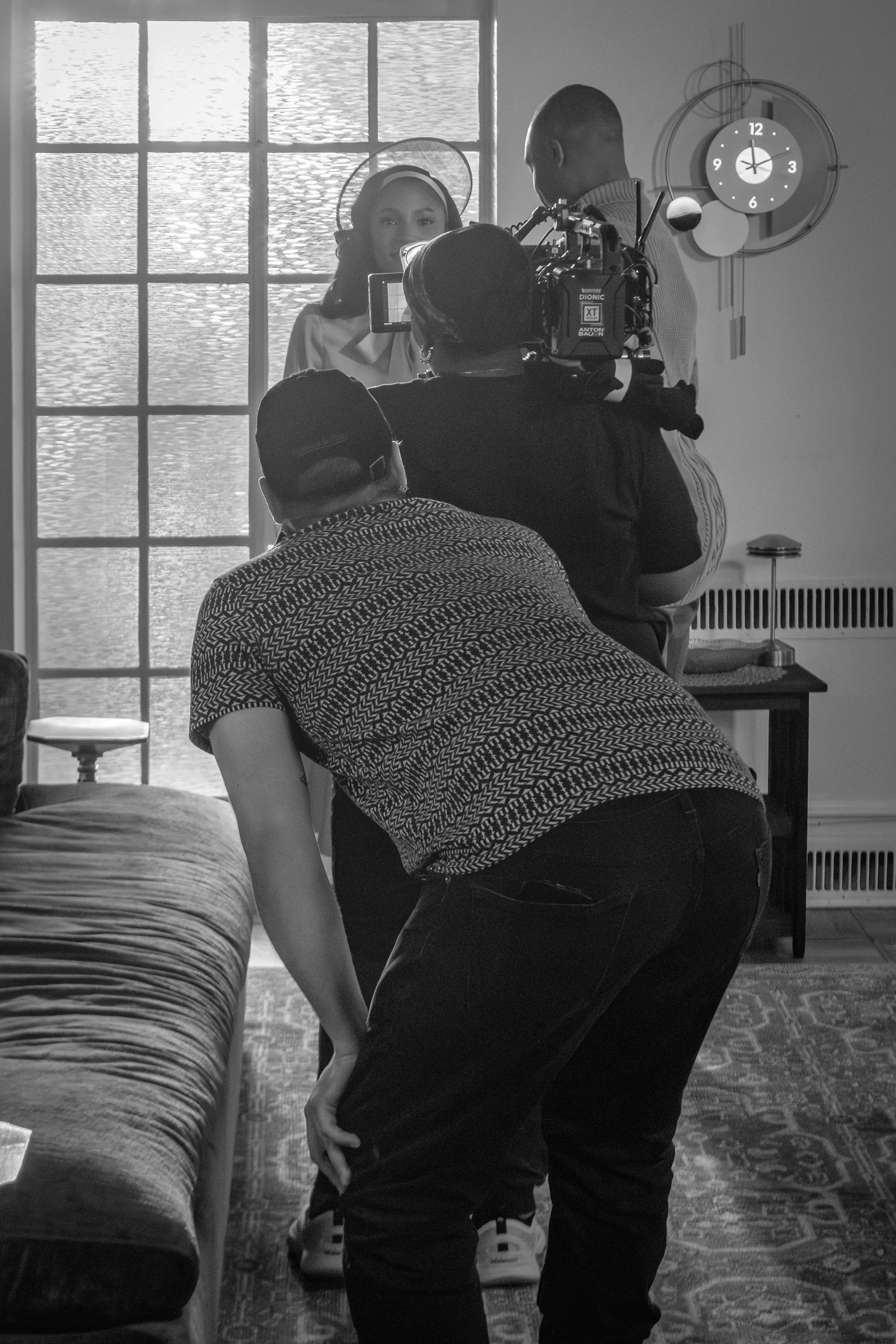 A person kneeling by a bed, making the bed. In the background, a woman is speaking to someone with a camera and a person with a camera is standing nearby taking a photo. The room has a window with curtains, a wall clock, and a patterned carpet.
