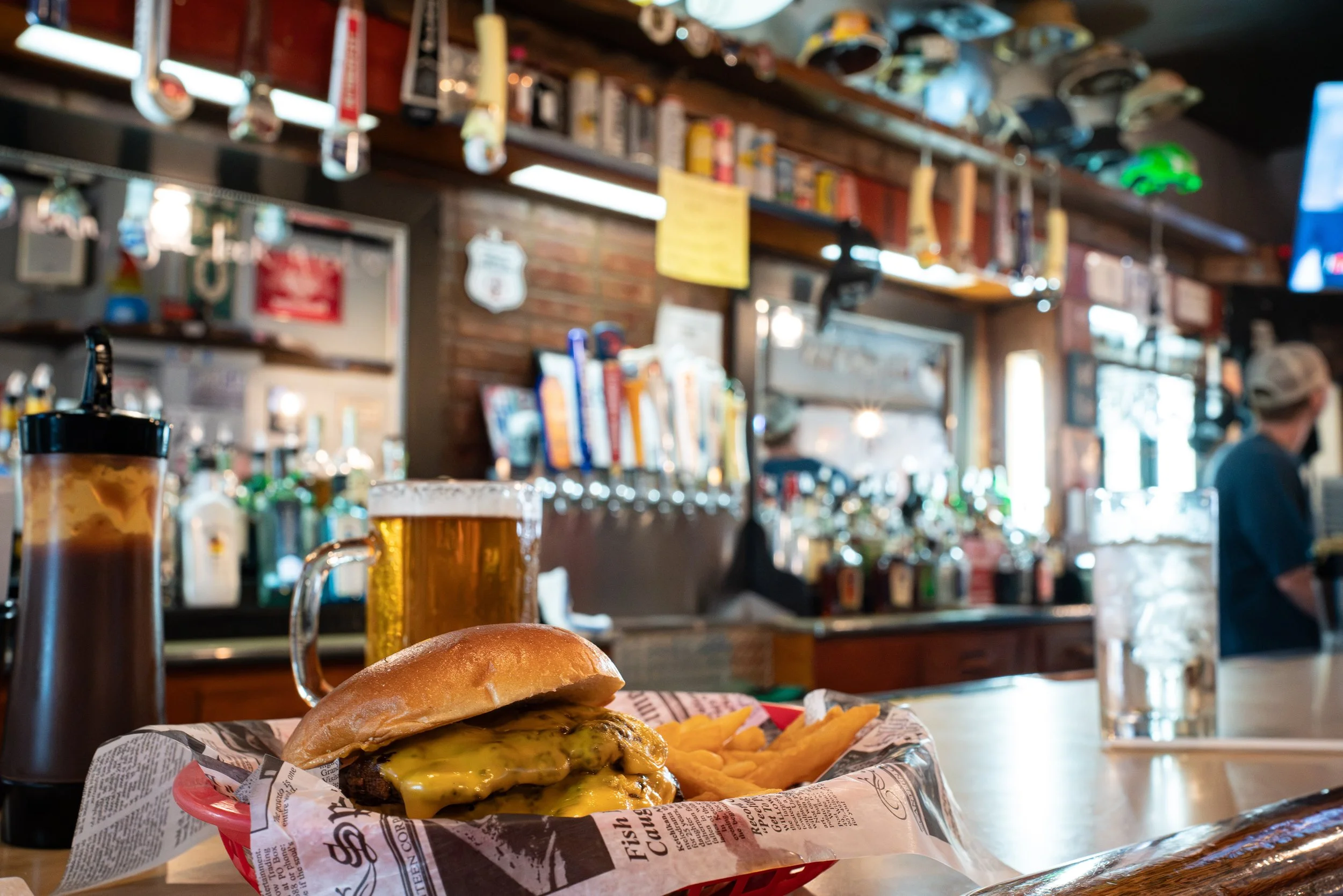 Close-up of a cheeseburger with melted cheese, pickles, and a bun in a basket lined with newspaper, alongside French fries, on a bar counter. In the background, a bar with liquor bottles and taps and a person wearing a cap are visible, with a blurred
