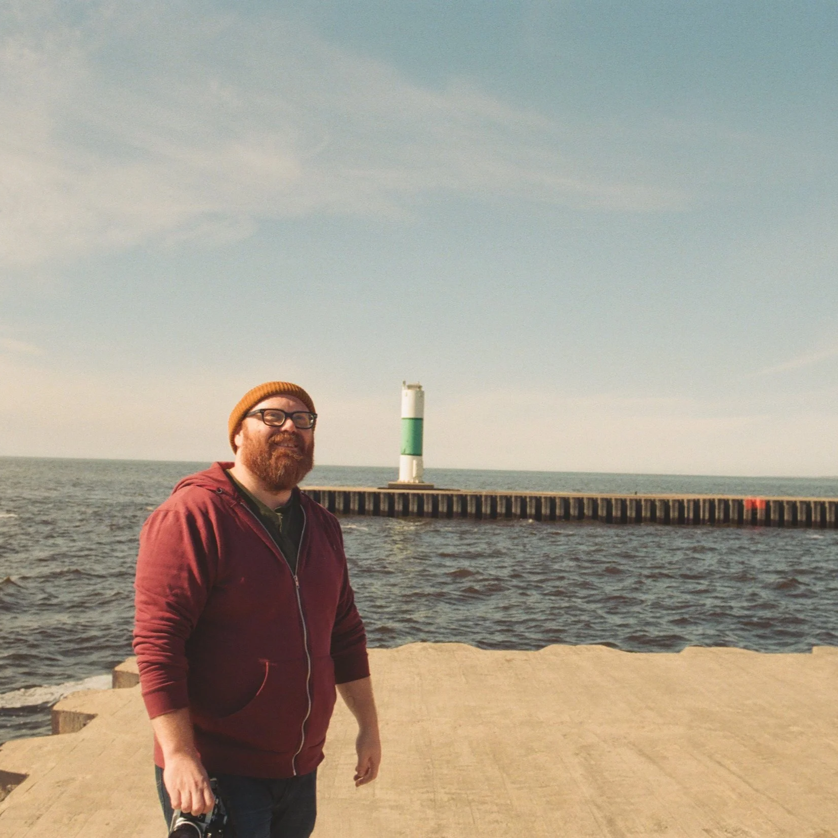 A man wearing a maroon hoodie, glasses, and a brown knit cap stands near a body of water on a pier, with a green and white lighthouse in the background under a partly cloudy sky.