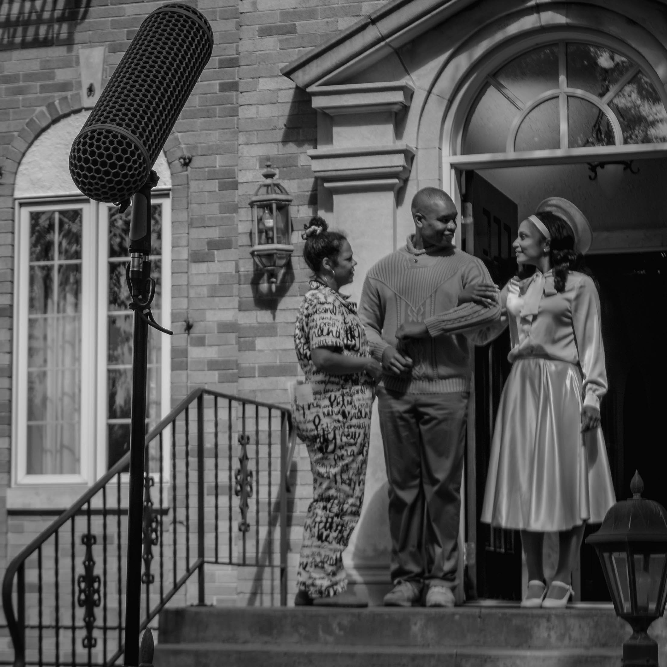 Black and white photo of three women standing on outdoor stairs. One woman is wearing a patterned dress, the second woman is wearing a sweater, and the third woman is wearing a dress with a headband. They are in front of a brick house with a large ar