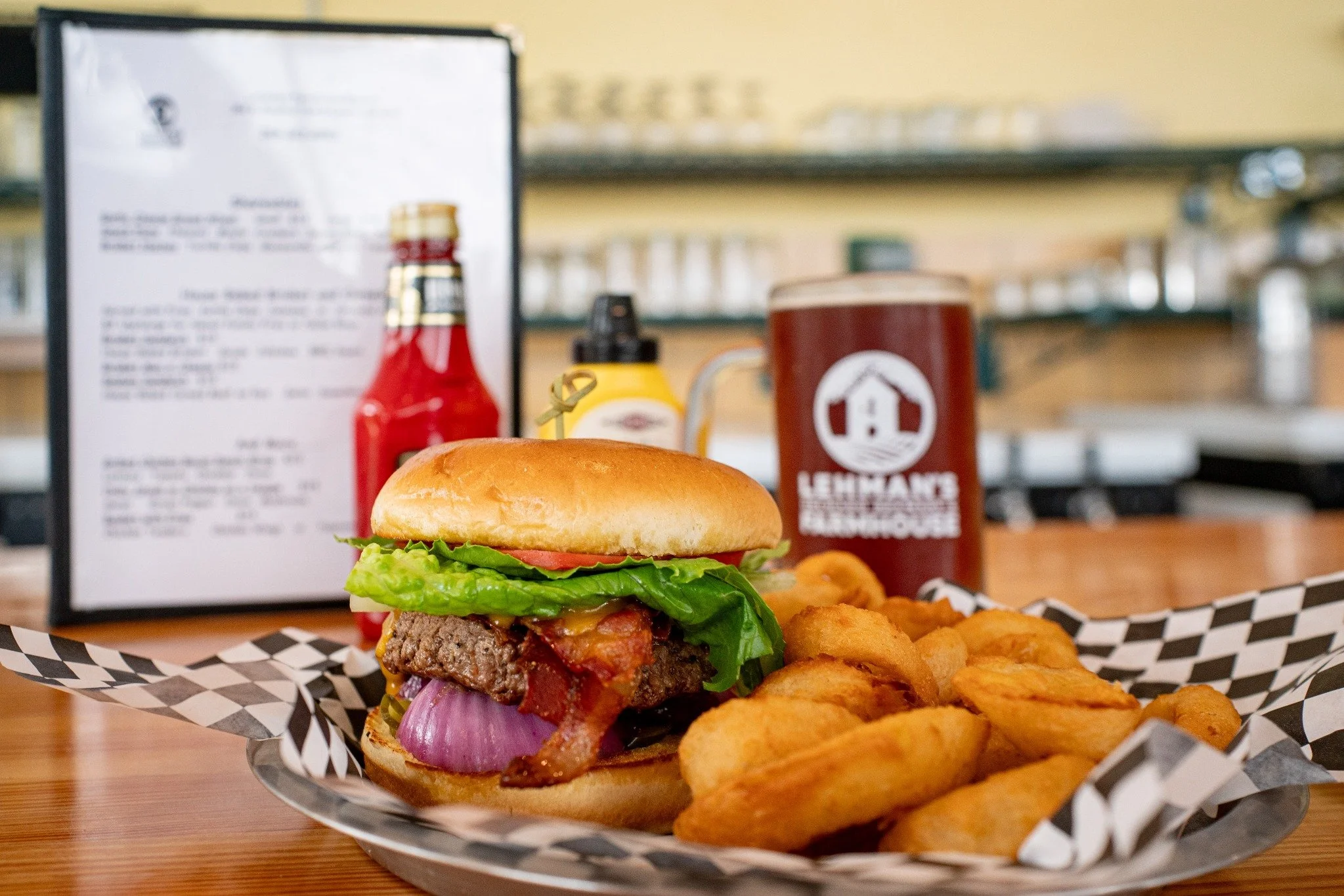 A cheeseburger with lettuce, tomato, onion, bacon, and a toasted bun, served with a side of seasoned potato wedges on black and white checkered paper at a diner-like setting.