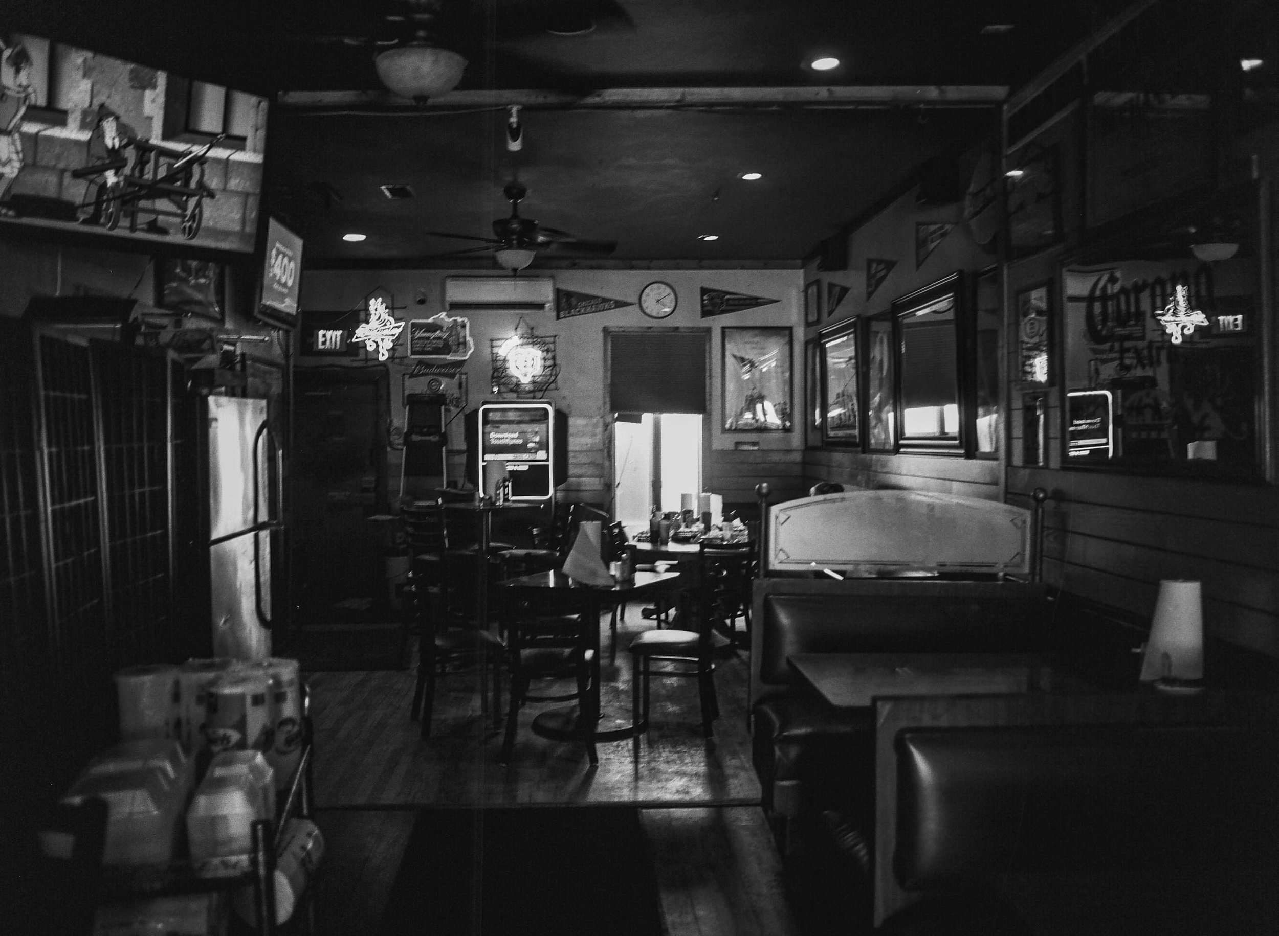 Interior of a dimly lit restaurant or bar with tables and chairs, neon signs on the walls, and a clock showing approximately 8:25.