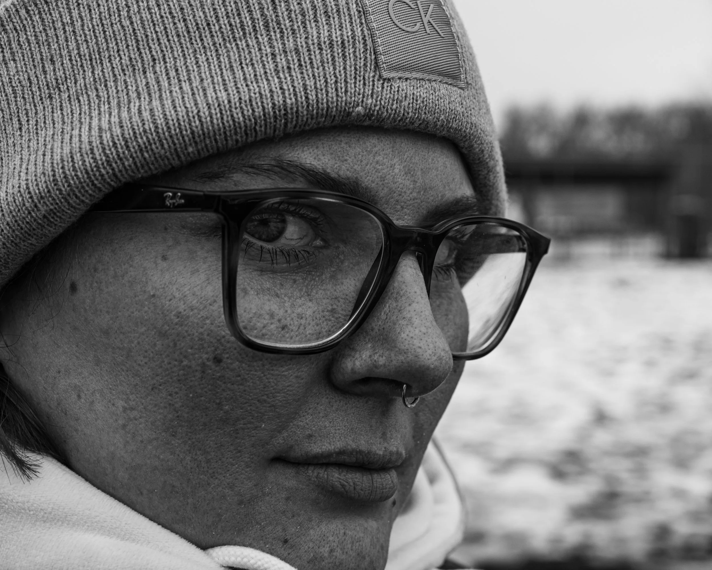 Close-up of a person's face wearing glasses and a knit beanie cap, looking down with a blurred background.