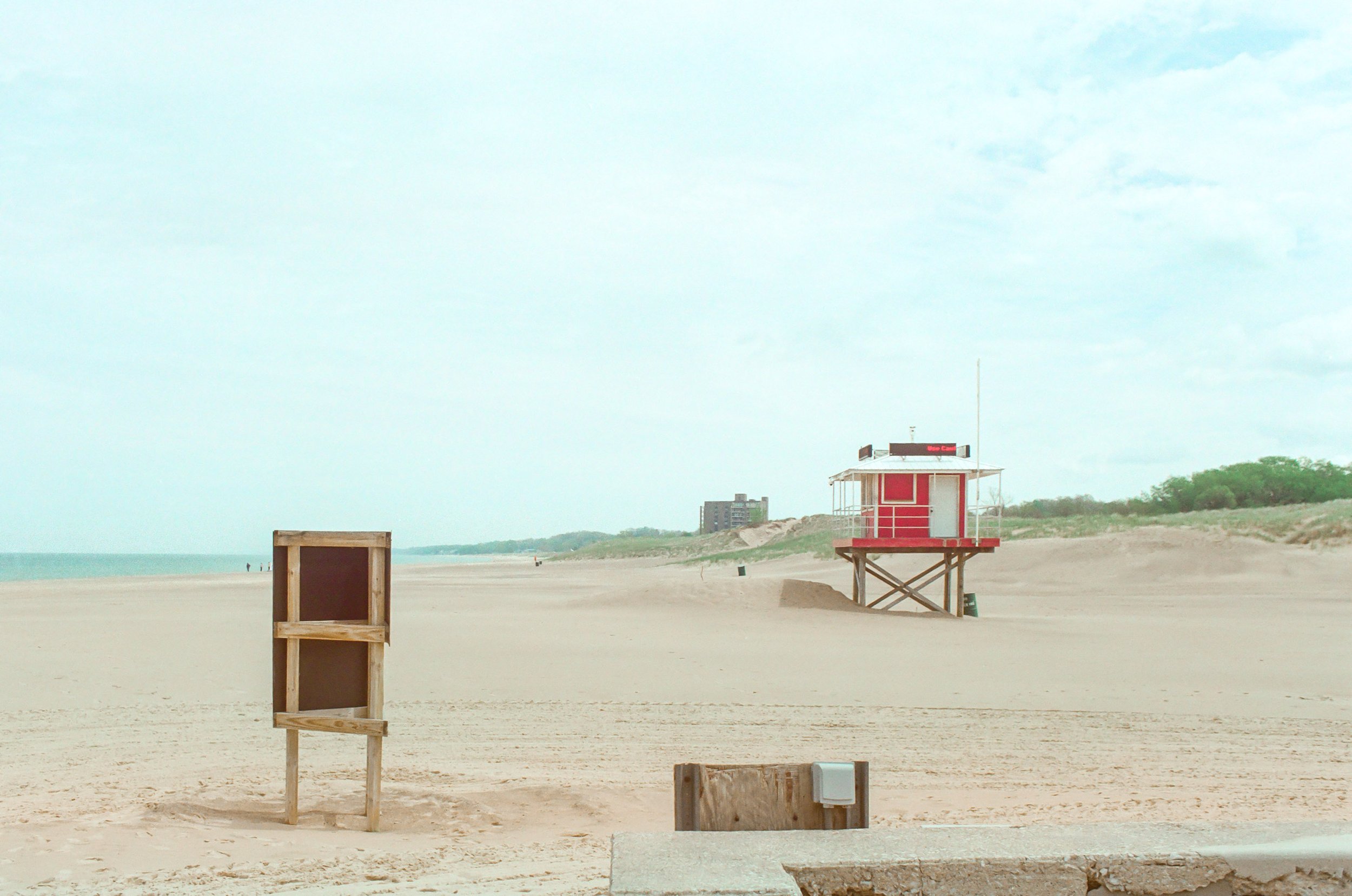Empty beach with wooden lifeguard stand, small wooden box near beach, and a small building in the distance with greenery on the horizon under a cloudy sky.