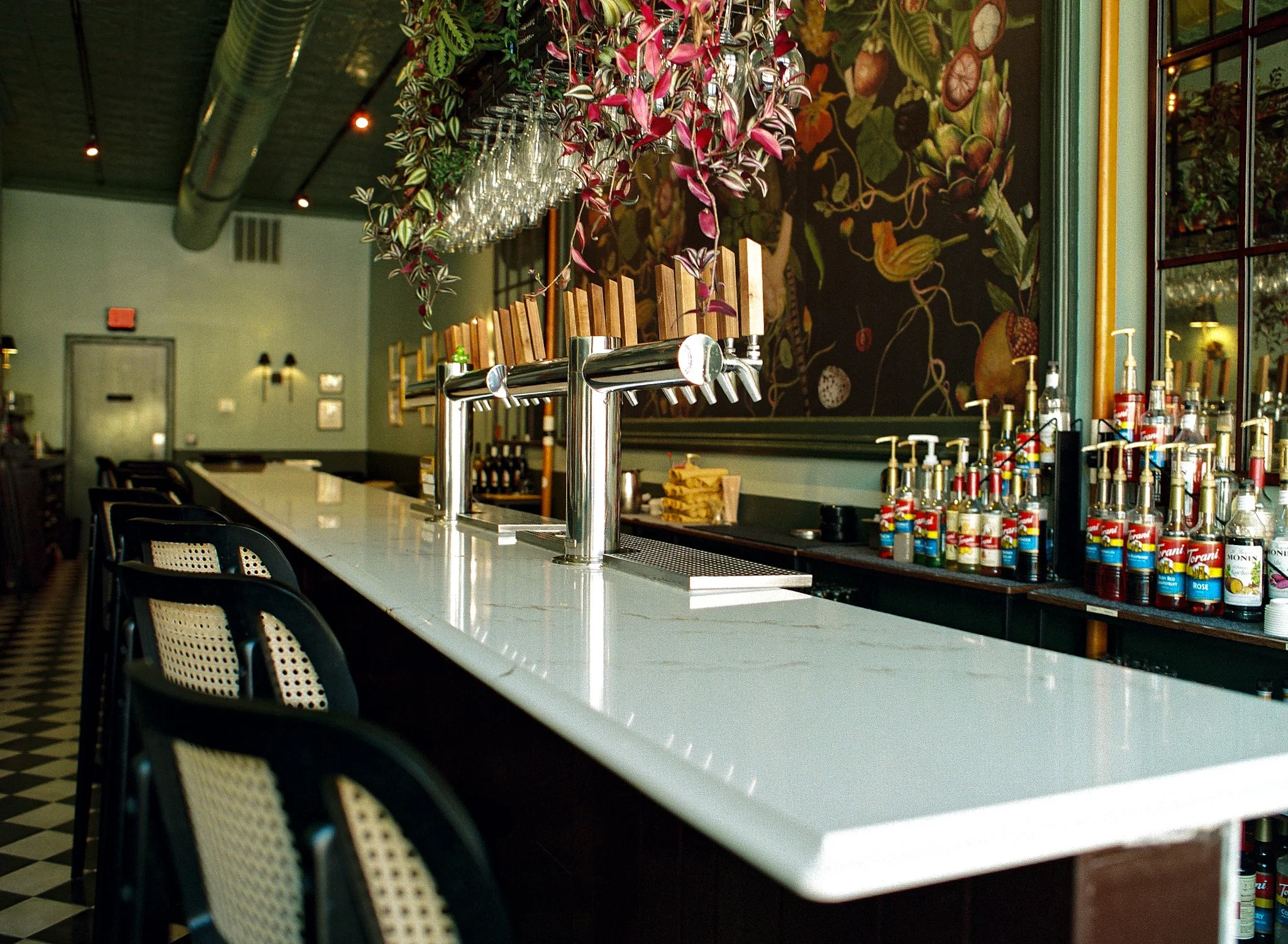 Interior of a bar or restaurant with a long white counter, black chairs with woven seats, a hanging plant, and a mural with fruits and vines on the wall. Various bottles and condiments are on the counter.