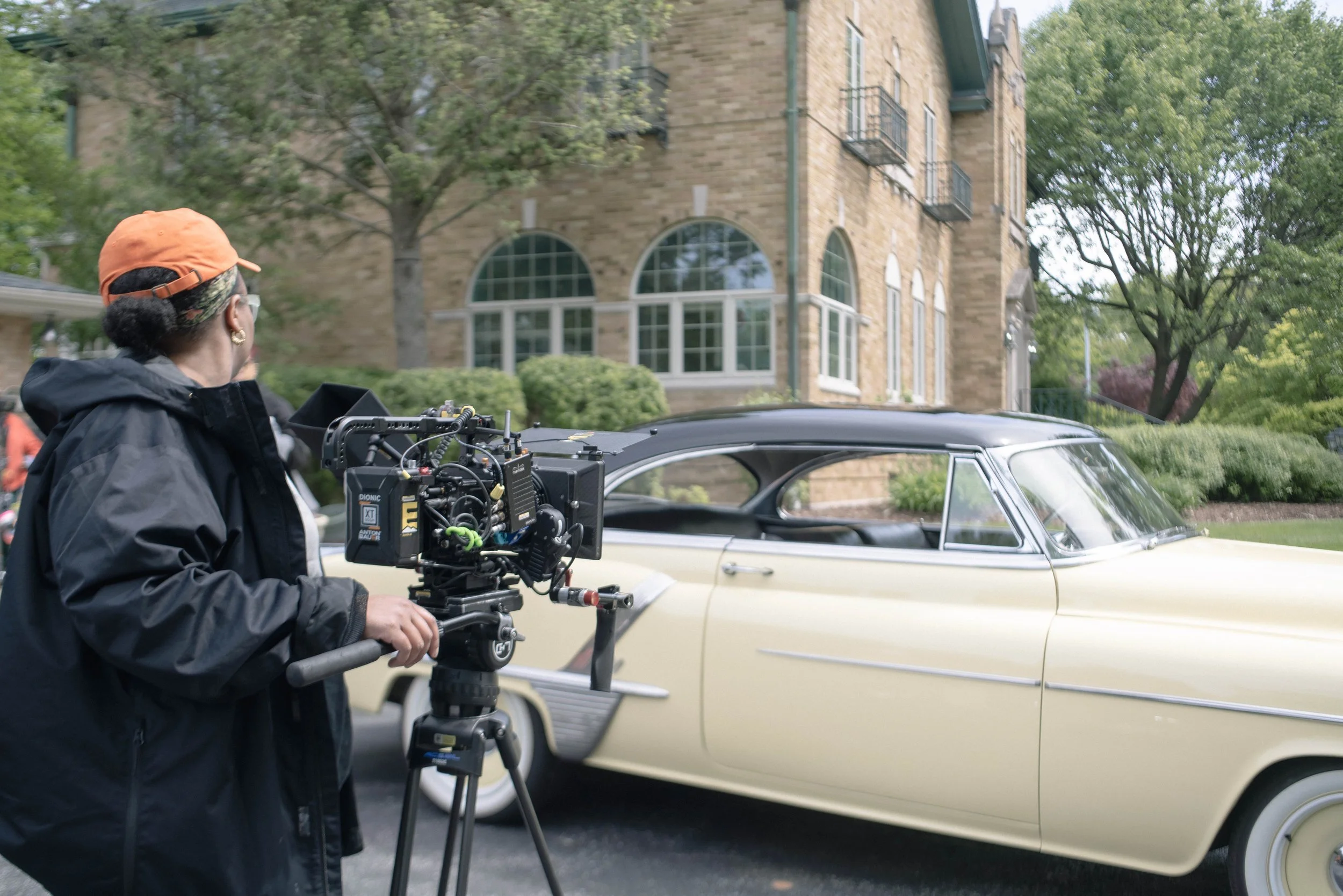 A woman operating a professional camera on a tripod in front of a vintage cream-colored car parked on a street, with a multi-story brick building and trees in the background.