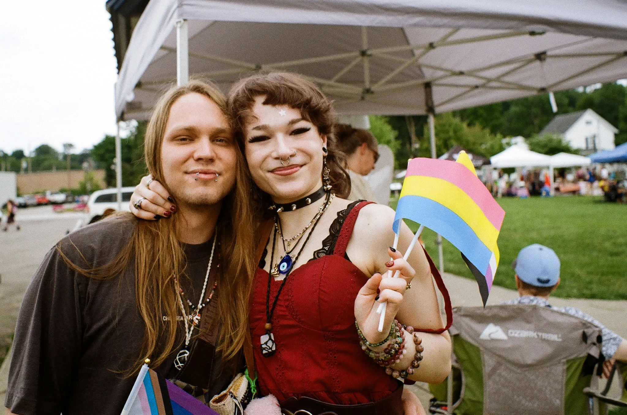 Two young women standing close together at an outdoor event, smiling at the camera. One is wearing a black shirt and has long, straight hair, while the other has short, curly hair with star-shaped face stickers and is holding a rainbow-colored pinwhe