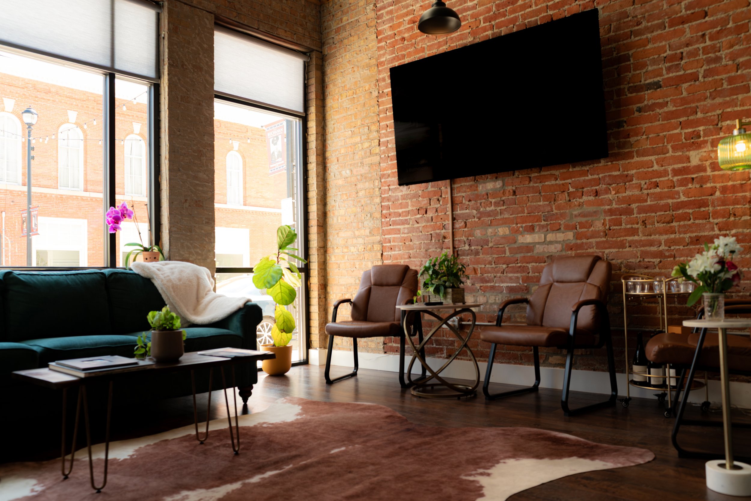 A cozy living room with large windows, exposed brick walls, a black sofa with white pillows, potted plants, a coffee table, and brown armchairs, with a wall-mounted television and a colorful hanging lamp.
