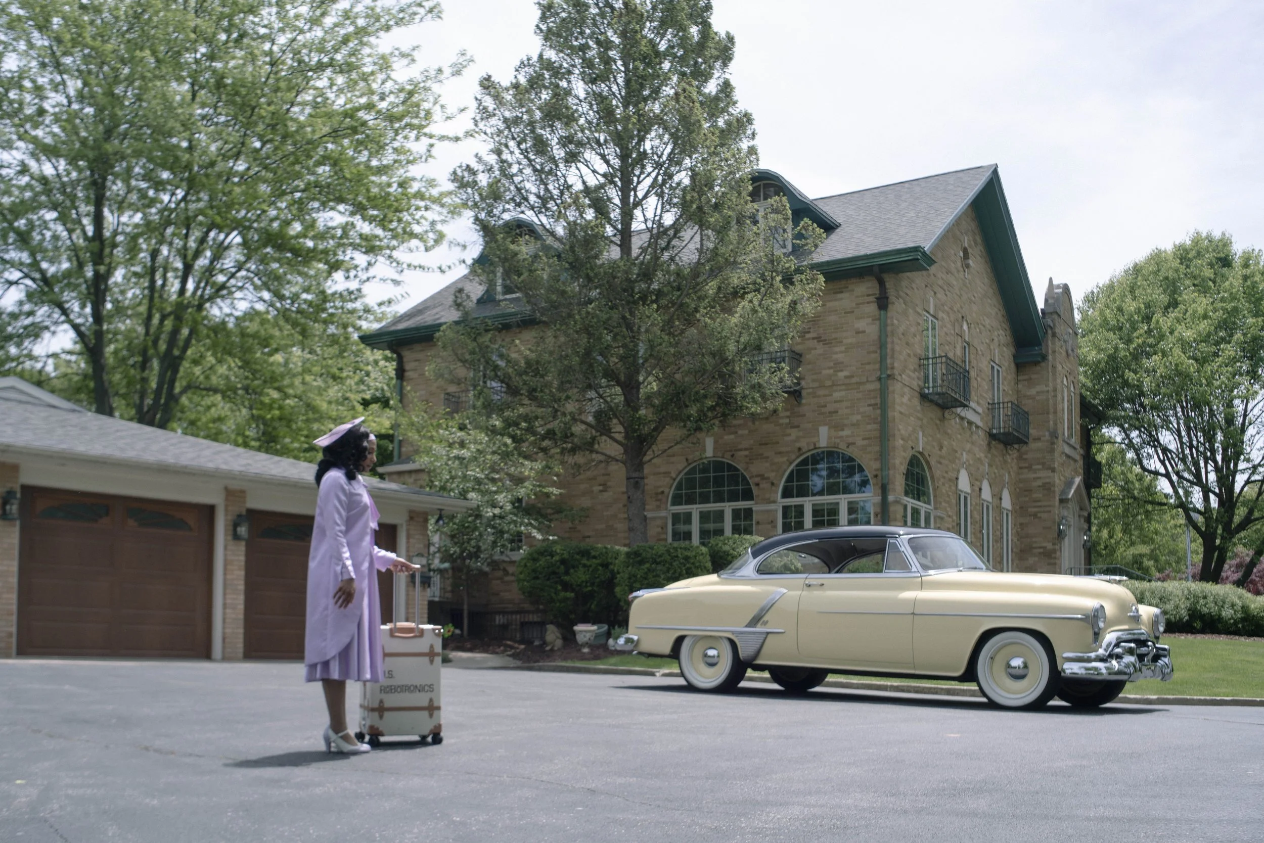 A woman wearing a light purple coat and white high heels, pushing a vintage suitcase on wheels, standing outside a large stone house with arched windows. The house has several stories with a steep roof and small balconies. A classic cream-colored car