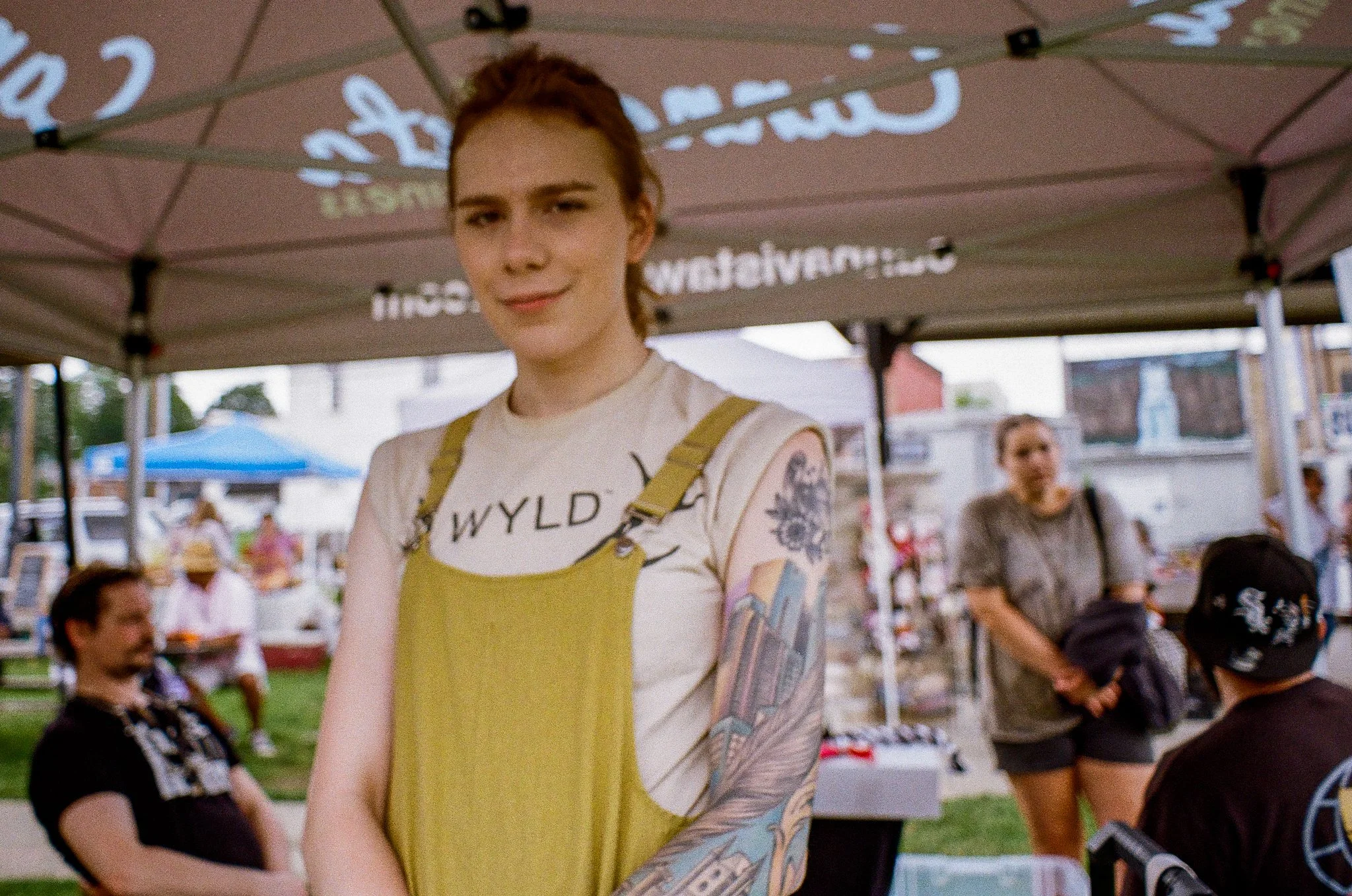 A young woman with red hair pulled back, wearing a beige T-shirt with black lettering and a mustard yellow apron, at an outdoor event under a tent with various people in the background.