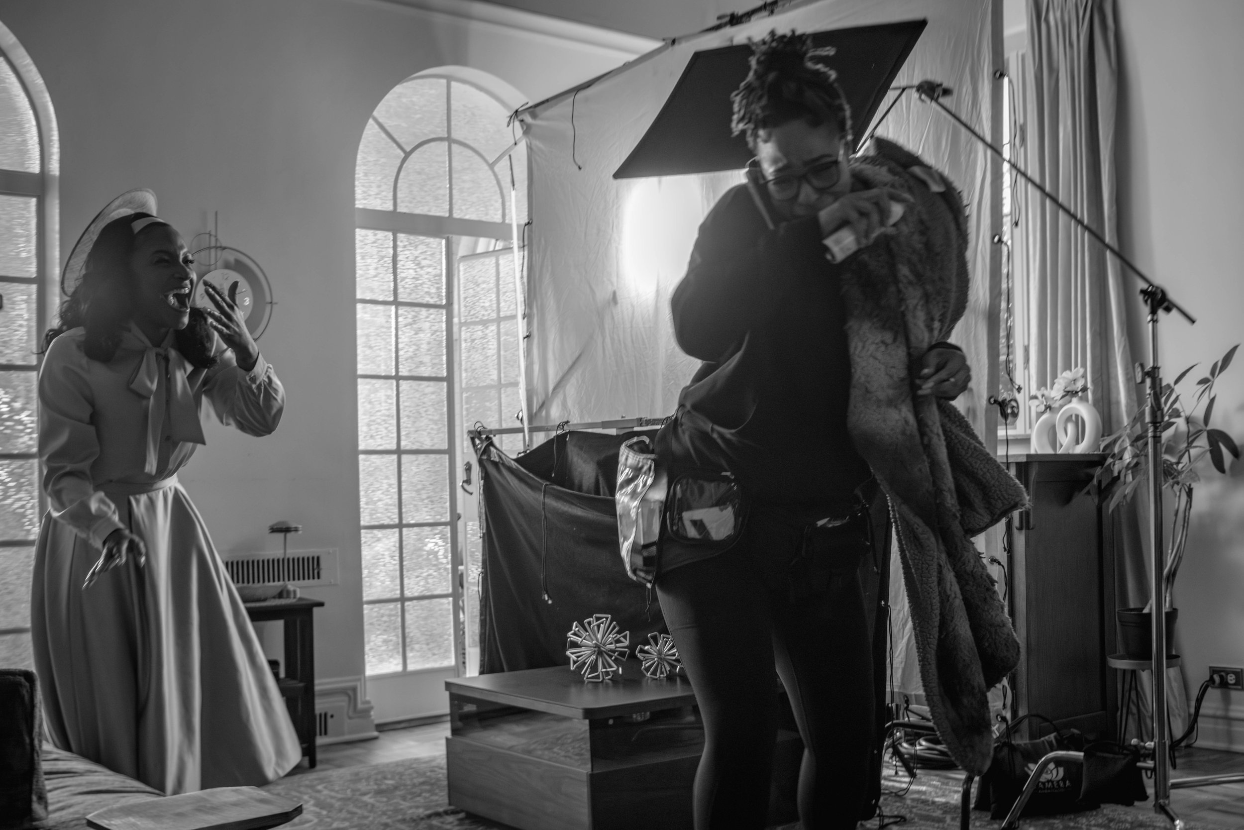 Black and white photo of a woman smiling and wearing a vintage dress with a wide-brimmed hat, standing near a table with a geometric sculpture. In the background, a man is holding a camera gear, and studio equipment is visible, with large windows and