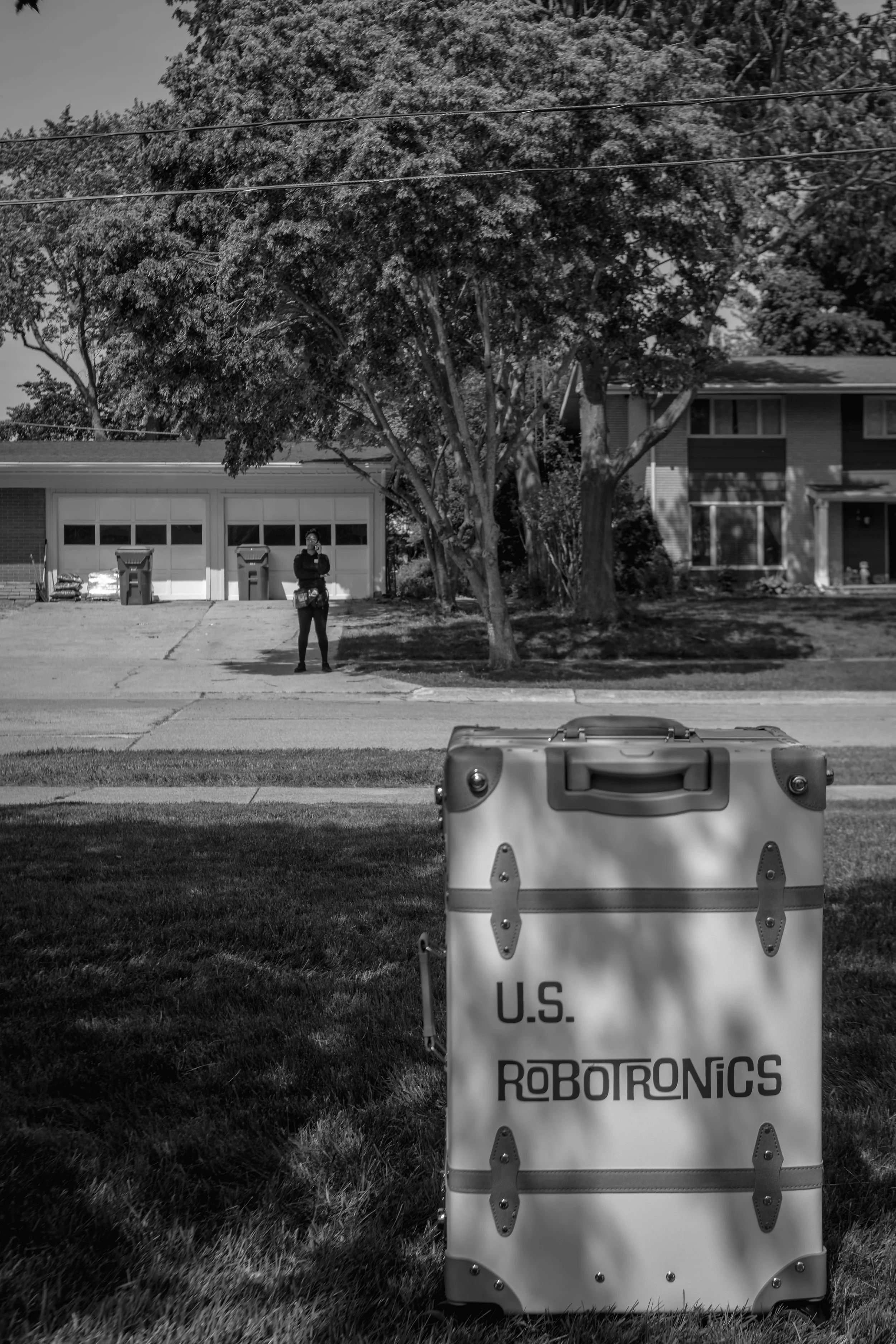 A person standing in front of a house with trees and a garage, holding a device to their ear. In the foreground, there is a large container labeled "U.S. ROBOTRONICS".
