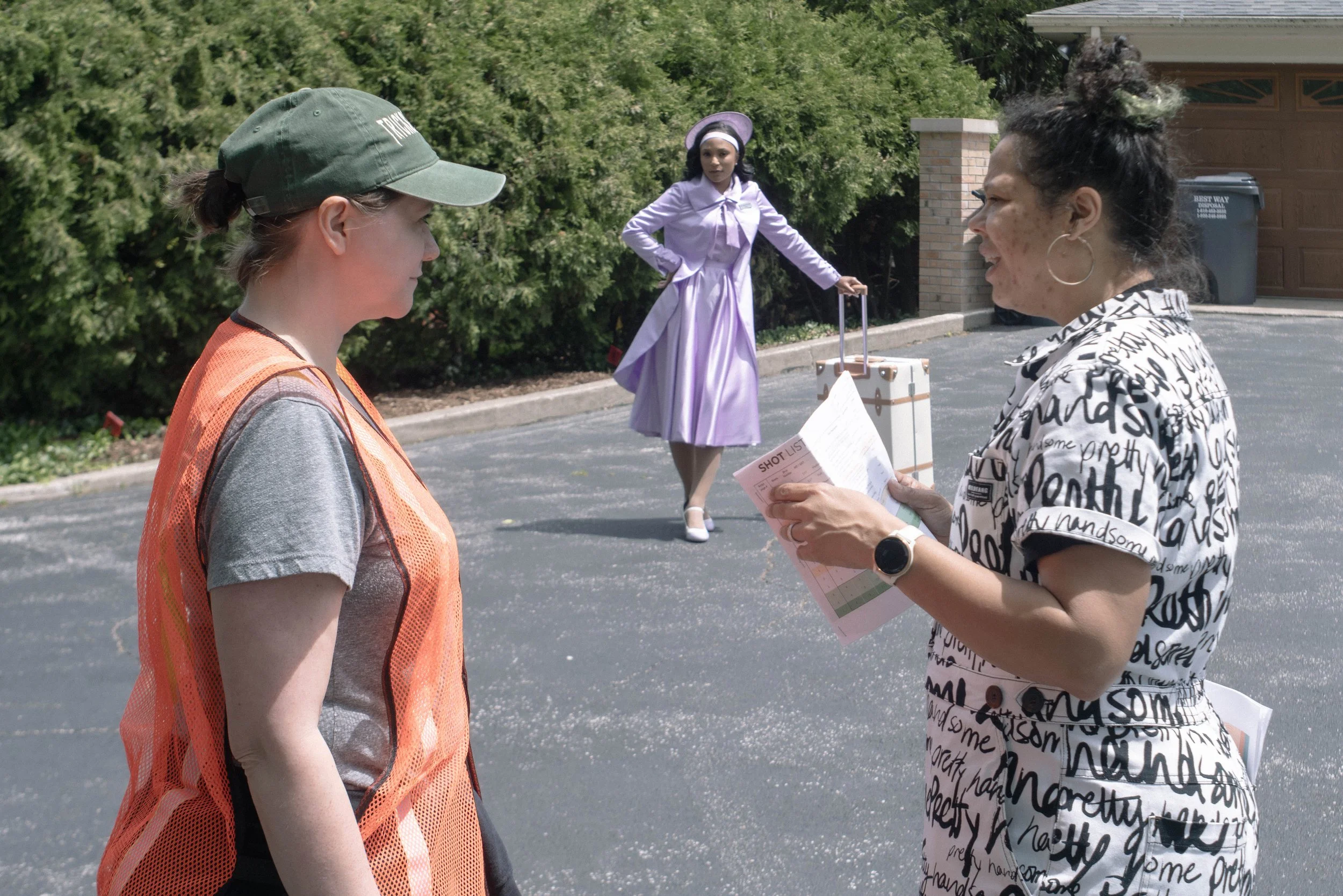 Two women are standing outdoors having a conversation, while a woman in vintage dress and hat with a suitcase stands in the background on a street with greenery.