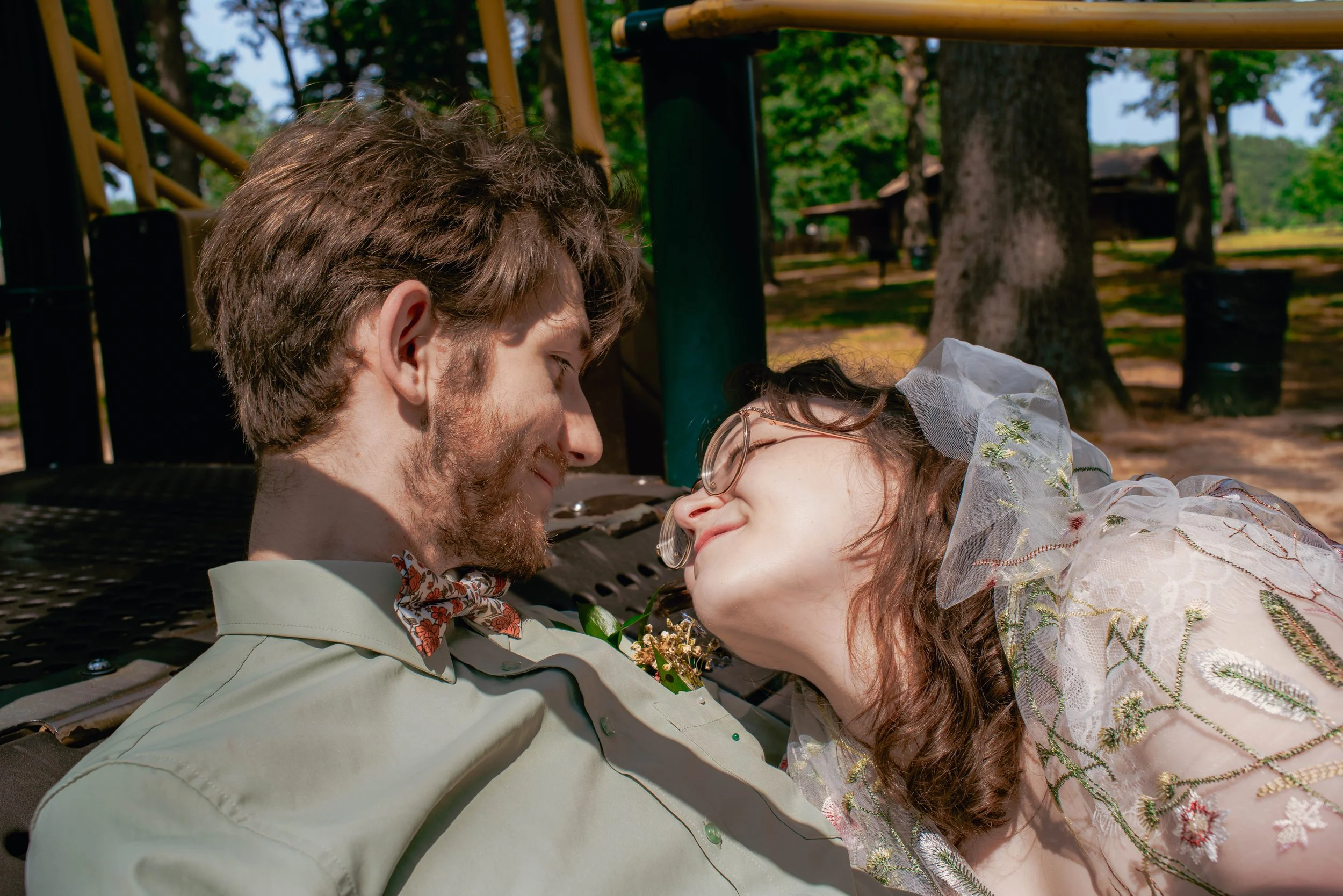 A man and a woman with closed eyes and smiling faces are lying on a bench outdoors, about to kiss. The man has brown hair and a beard, wearing a light-colored shirt and bow tie. The woman has long, brown hair, glasses, and is dressed in an embroidere