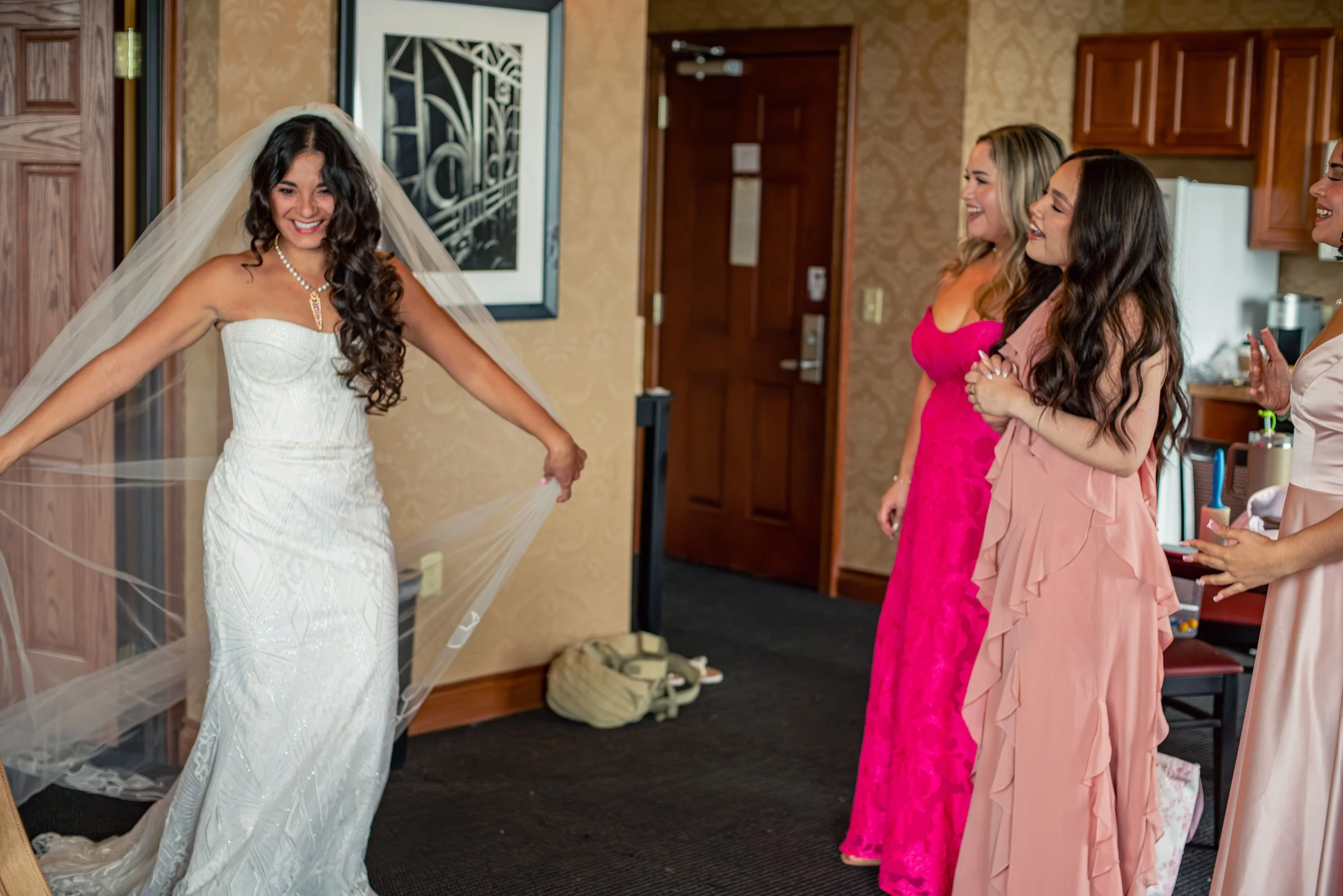 A bride in a white wedding dress and veil smiling as she stands with her arms out, surrounded by three women in pink dresses who are smiling and looking at her.