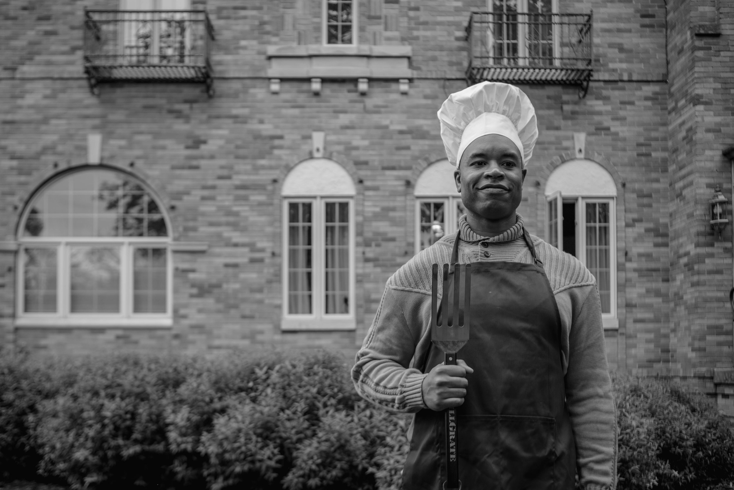 A man in a chef's hat and apron holding a spatula stands outdoors in front of a brick building with multiple windows.