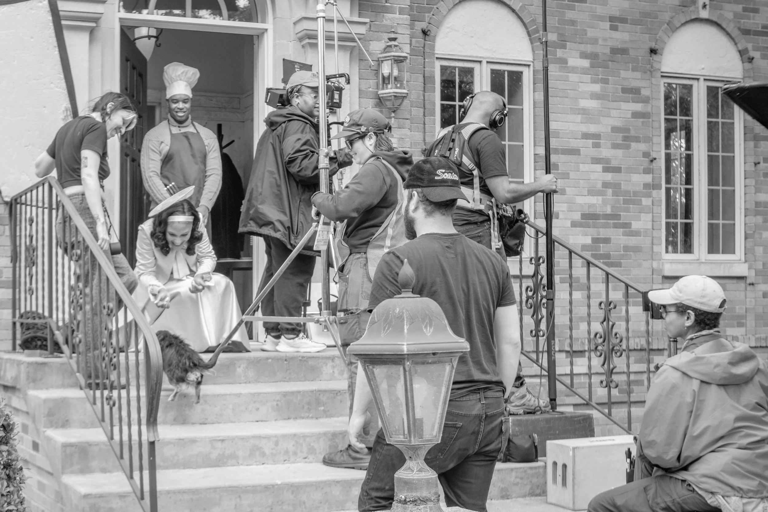 Children and adults filming a scene on the front porch of a brick house. Two girls are seated on the steps, one wearing a hat and the other in a medical mask. One woman in a chef hat stands near the door, while another person with headphones and a ba
