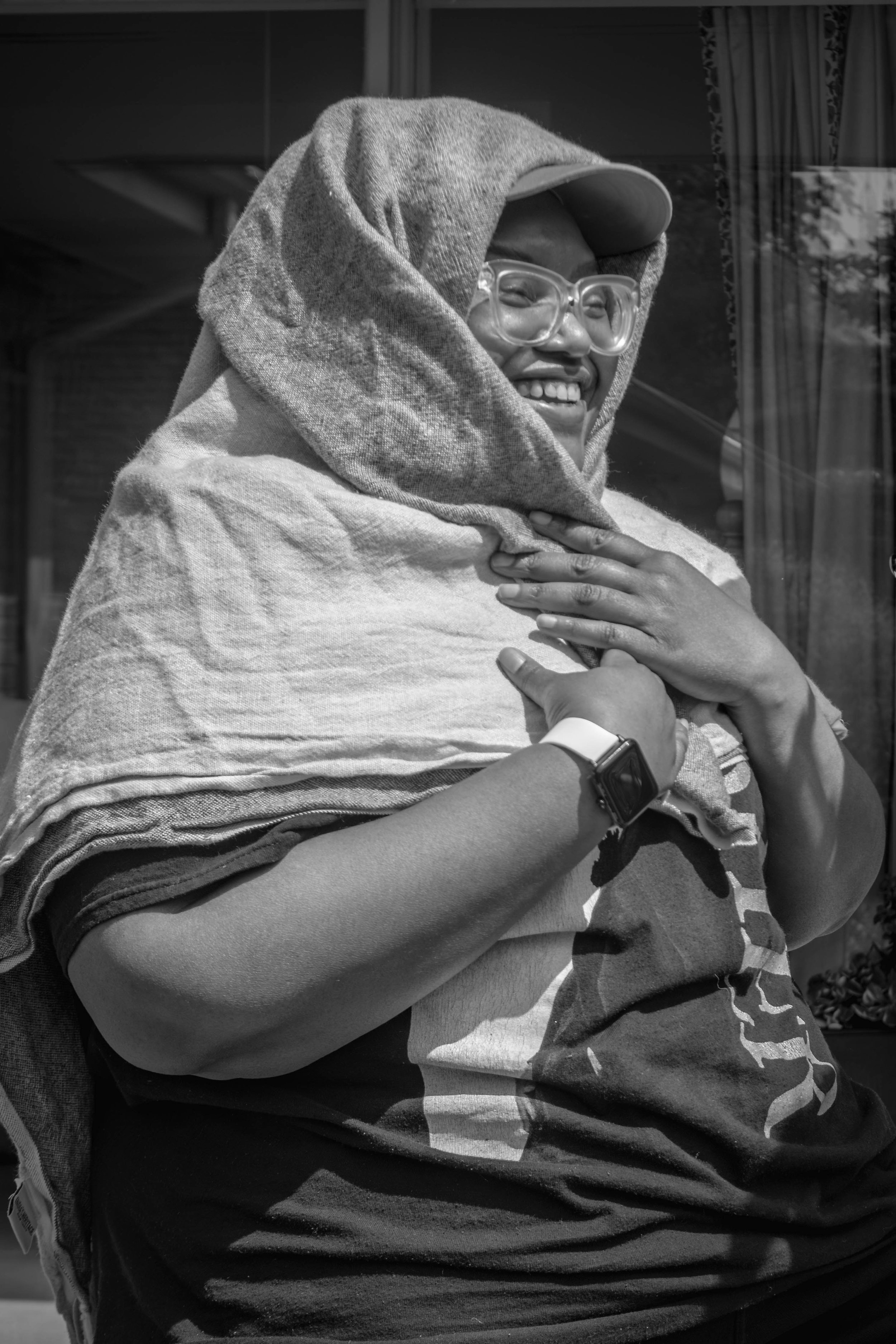 A person smiling, wearing glasses, a cap, and a hoodie, bundle wrapped in a blanket, with an arm and hand showing a wristwatch, standing outside a window with curtains.