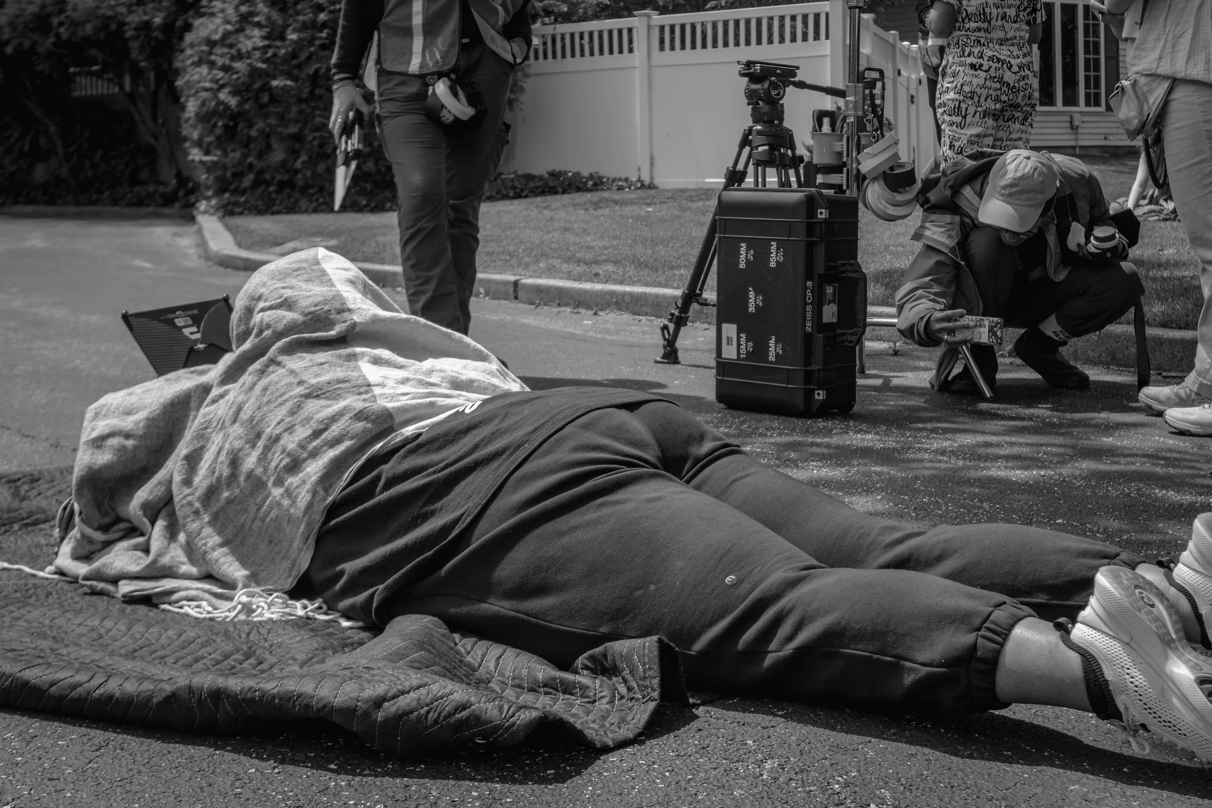Scene of a film or photo shoot on a street with a person lying on the ground covered with a blanket, surrounded by multiple people, camera equipment, and technical gear.