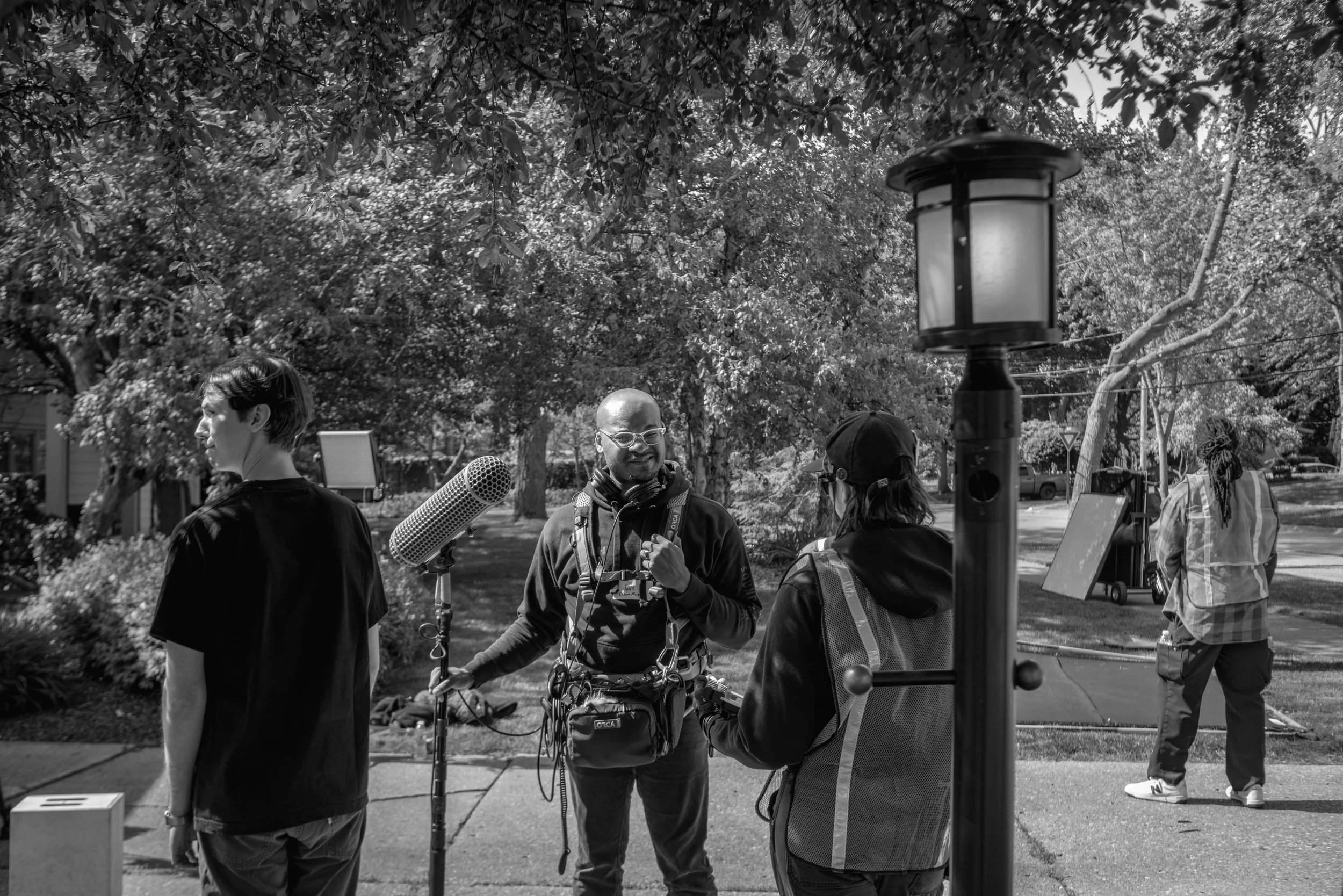 A group of people outdoors, with one person holding a microphone and others having conversations, surrounded by trees and a lamp post.