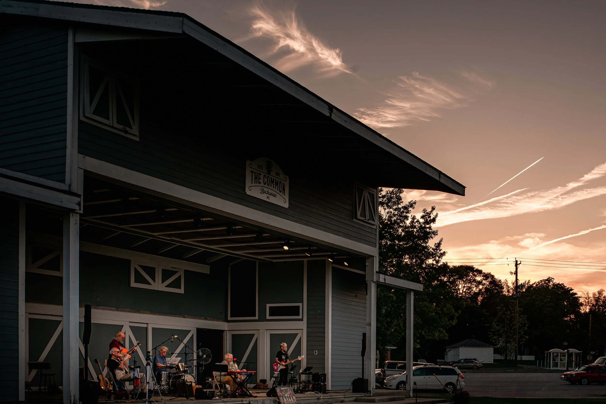 A live outdoor band performing on a stage at sunset with a sky full of clouds and contrails, attached to a small building labeled 'The Common' in Buchanan, Michigan, with people in the background and parked cars underneath.