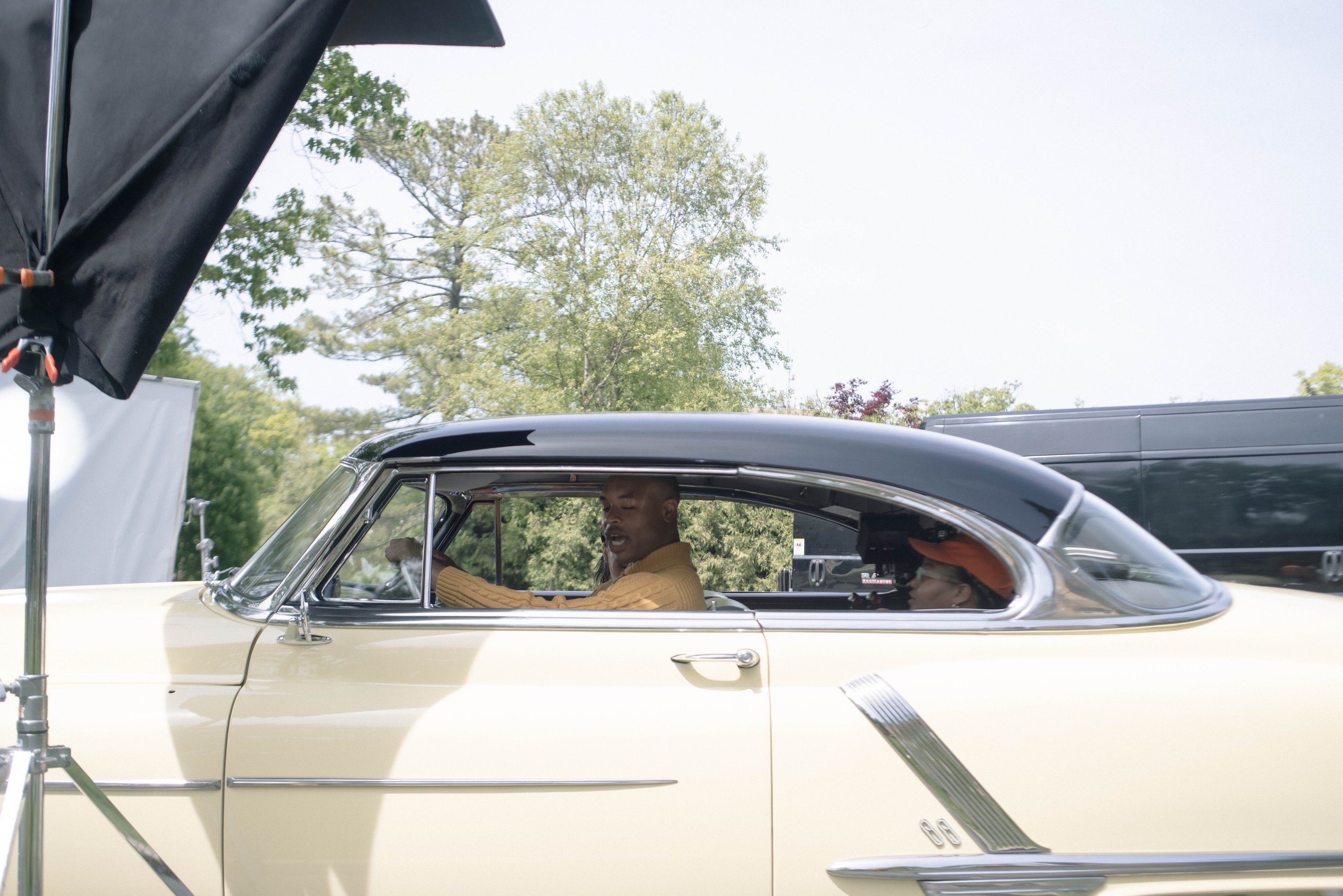 Two men inside a vintage car during a film shoot, with film set equipment and trees in the background.