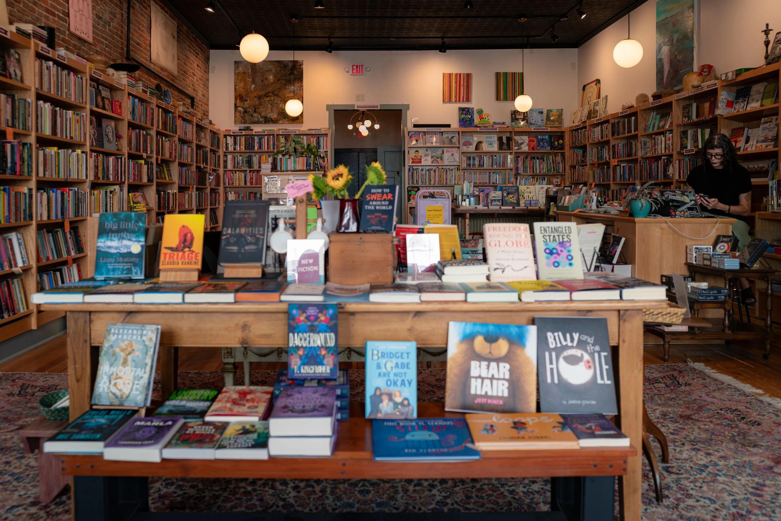 Interior of a bookstore with wooden shelves filled with books, a central table displaying more books, and walls decorated with abstract art and artwork, with a woman browsing on the right.