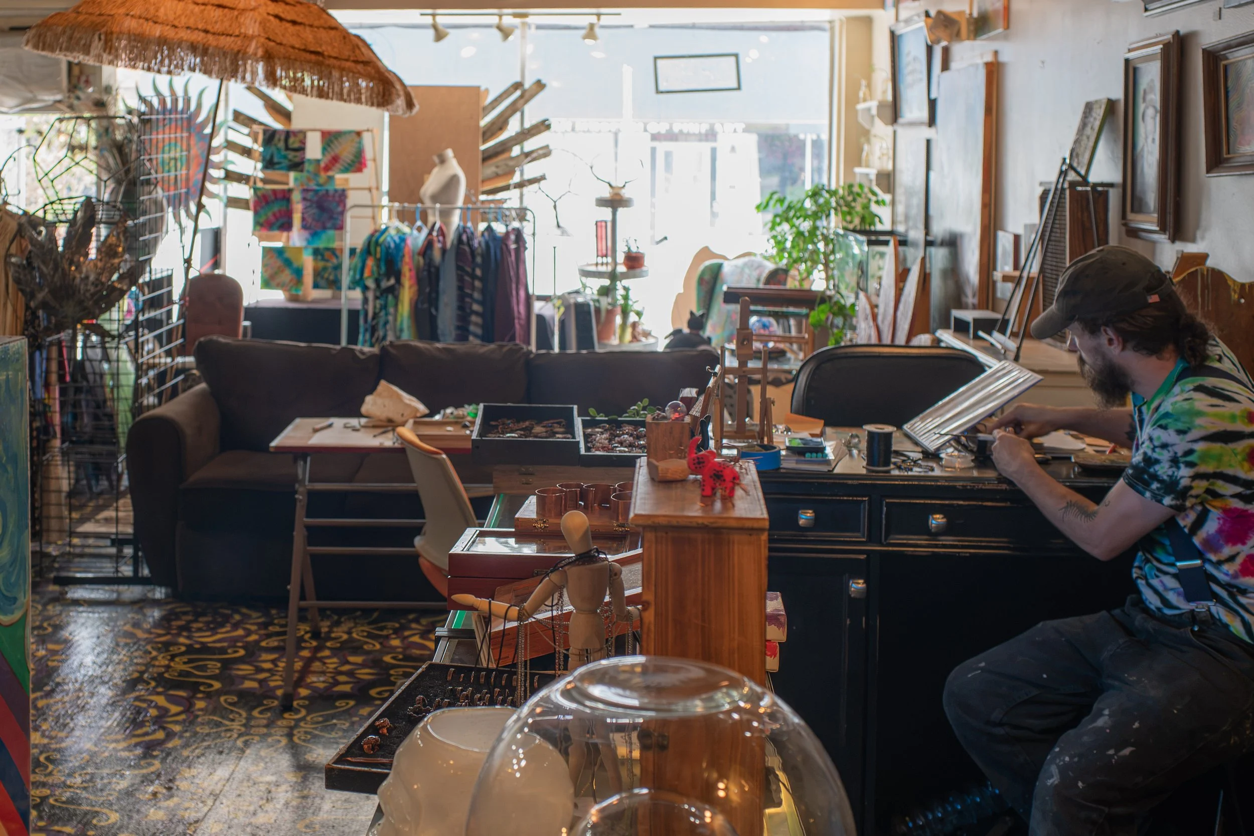 Inside a colorful thrift store with various clothing racks, paintings, and decorative items. A man is seated at a table working on jewelry or small crafts. Natural light streams through large front windows, illuminating the eclectic decor.