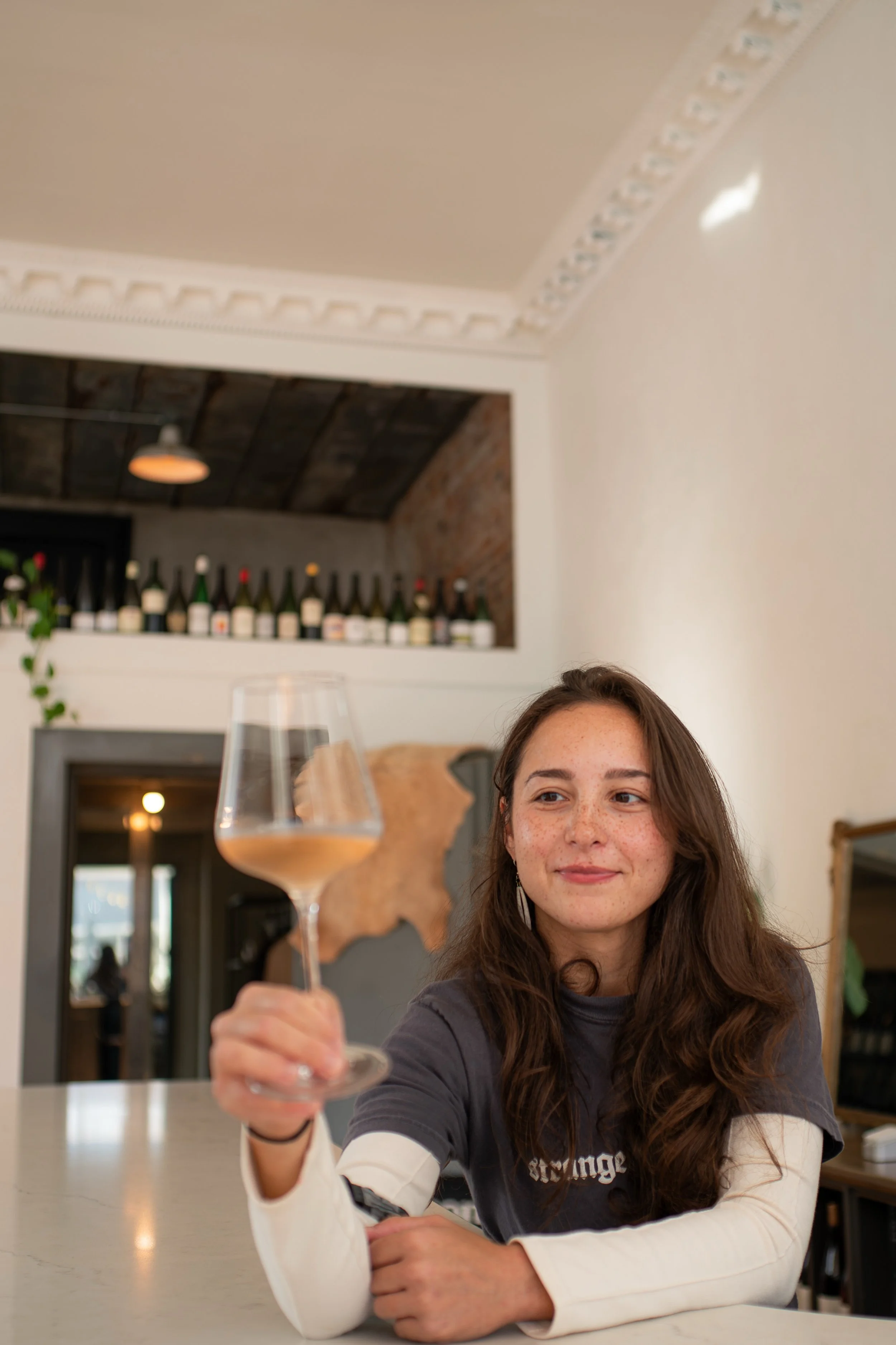 A young woman with long brown hair and freckles is sitting at a table, smiling, and holding a glass of wine in her outstretched hand. The background features a bar with wine bottles and a brick wall, with a rustic and cozy ambiance.