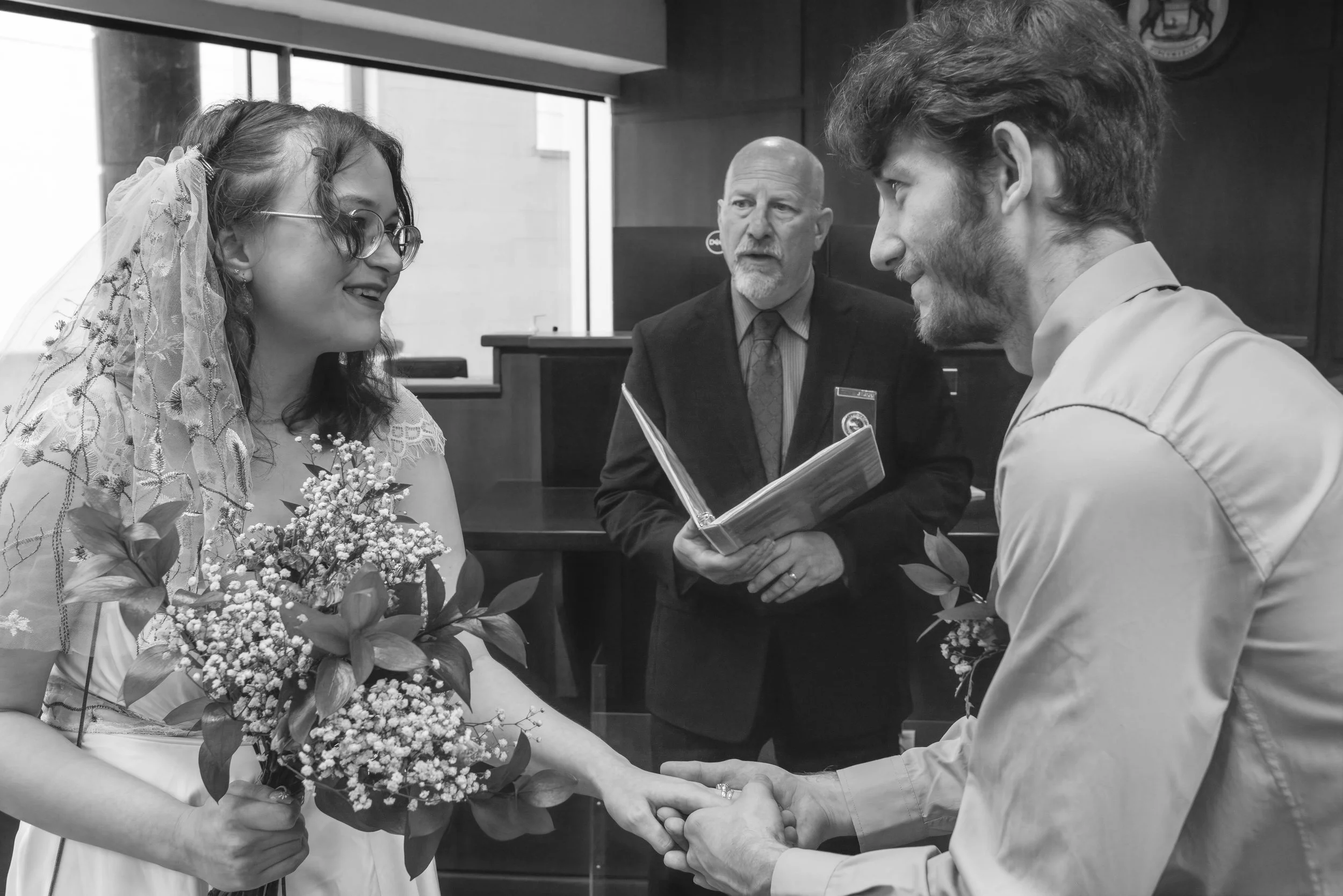A couple getting married, holding hands, with a priest officiating and another man reading from a book in the background.