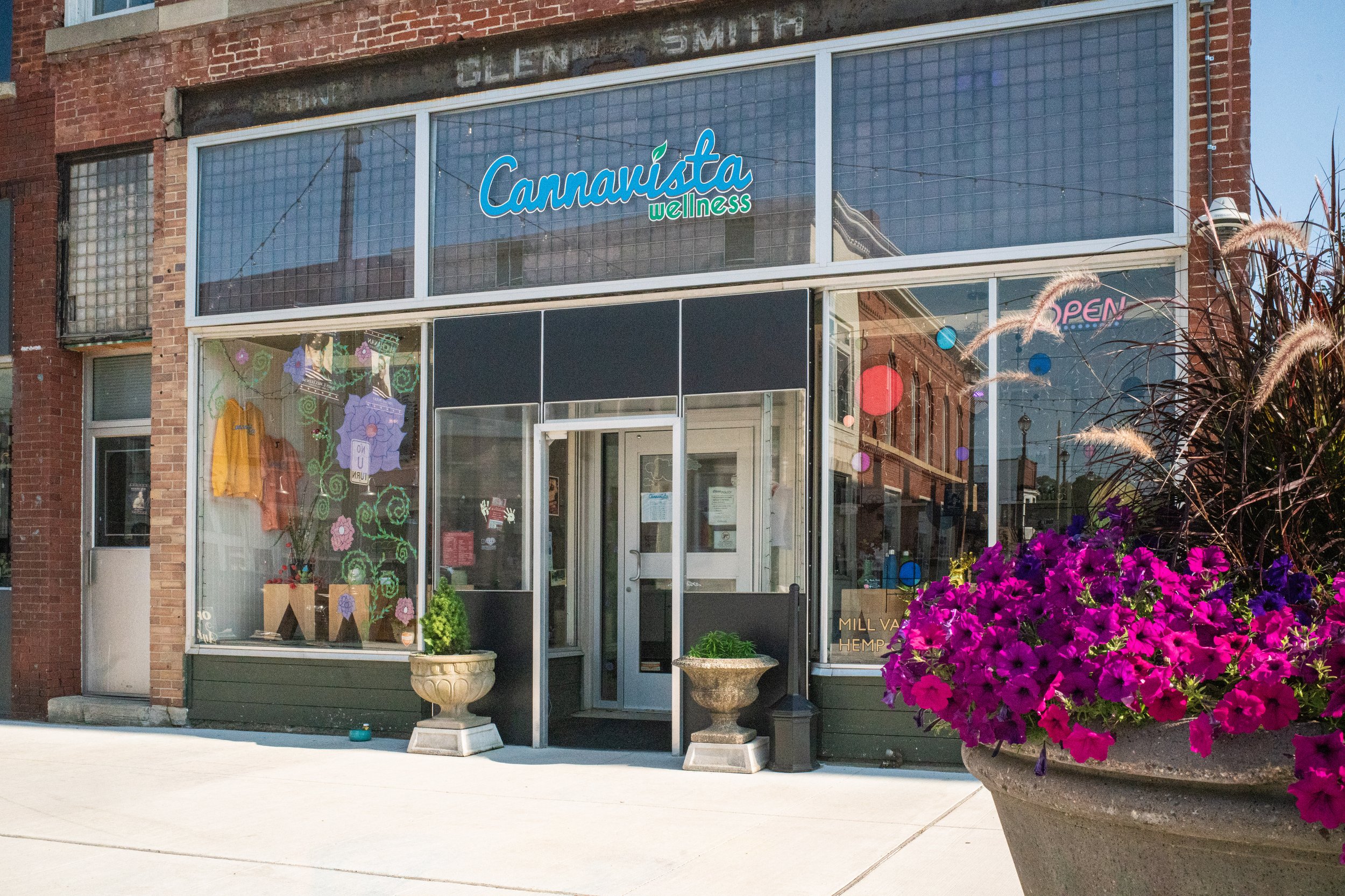 The storefront of a wellness shop named 'Cannaista Wellness' with large glass windows, colorful decorations inside, and vibrant pink and purple flowers in large planters outside.