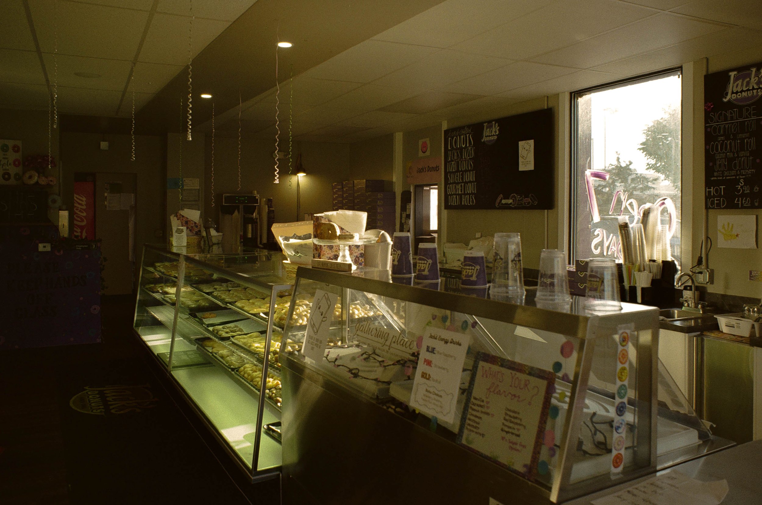 A display case filled with various baked goods inside a donut shop with menu boards on the wall and a window letting in natural light.