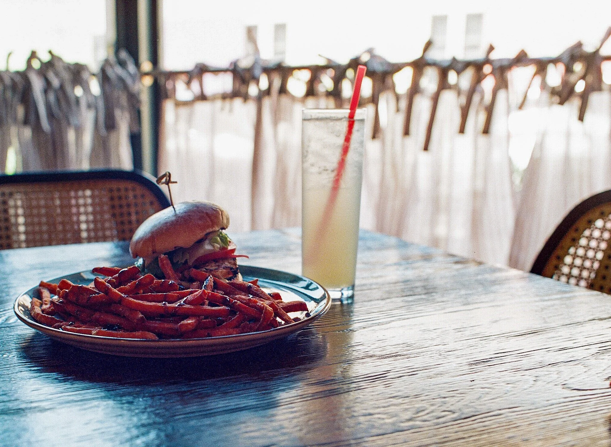 A plate of sweet potato fries on a wooden table with a burger and a drink in the background.