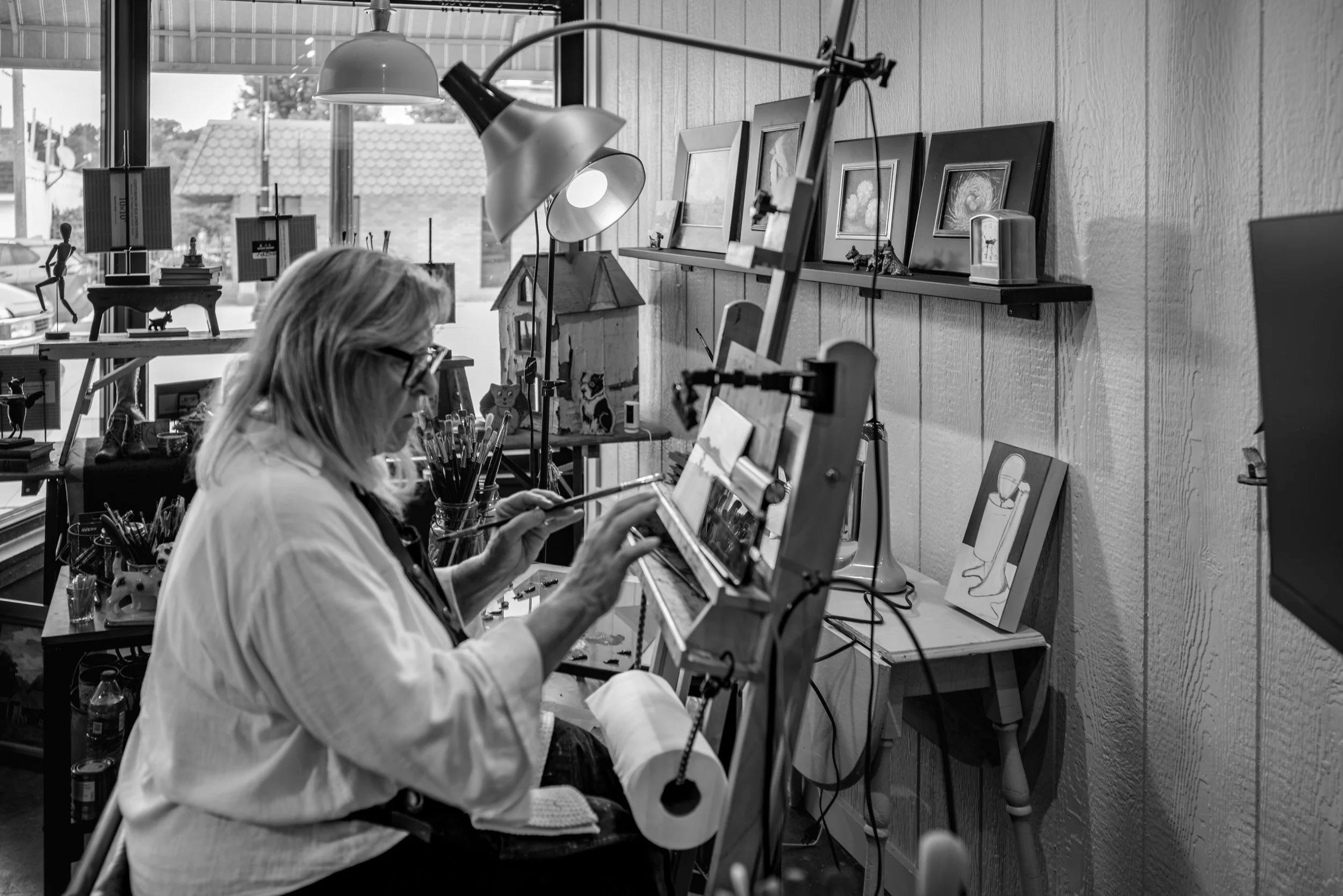A woman in glasses painting on a canvas in an art studio, surrounded by framed artwork, sculptures, and art supplies.