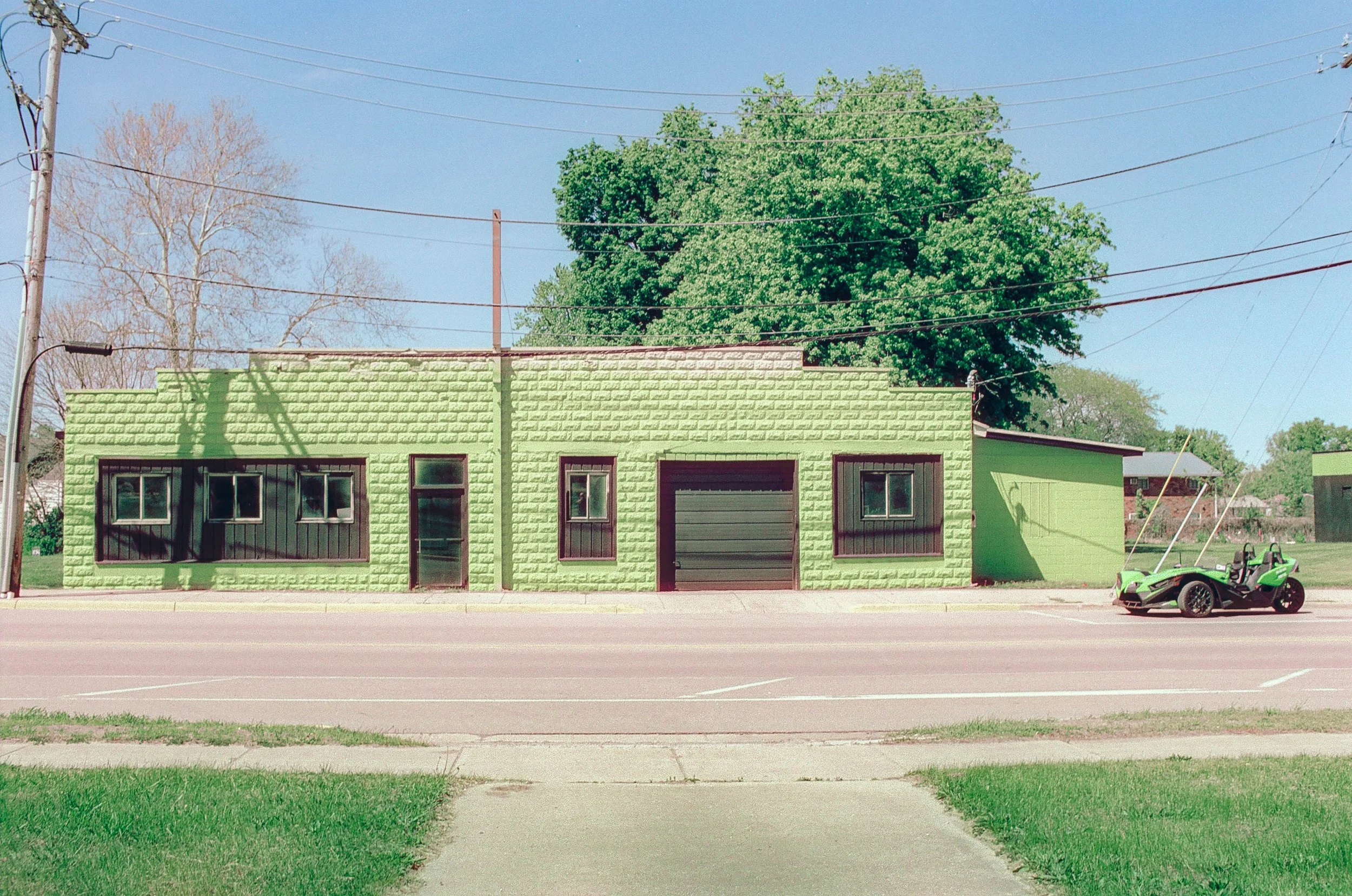A lime green brick building with four windows and a garage door, located on a street with a bike parked in front and a large green tree behind it.