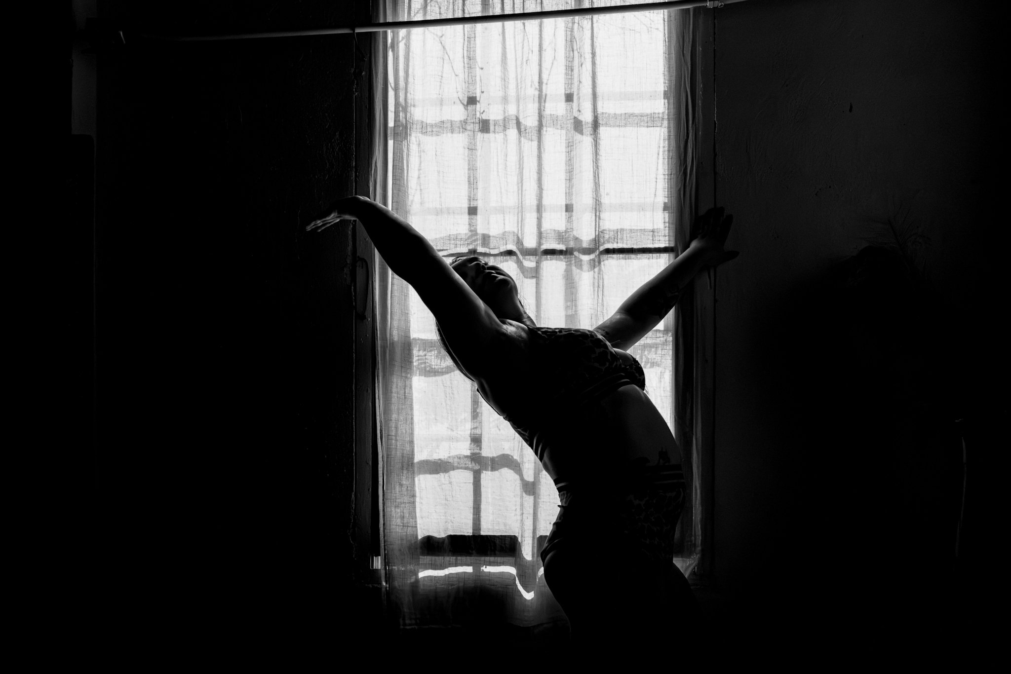Silhouette of a person stretching or dancing near a window with sheer curtains, black-and-white photo.
